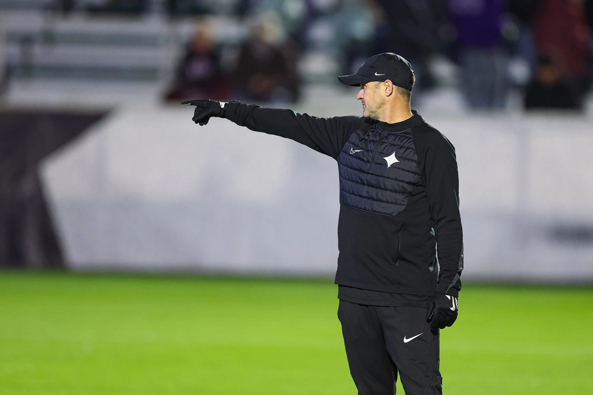A man wearing a black Nike cap and black sports jacket with a Nike logo, pointing with his right arm on the football field during a game or practice.