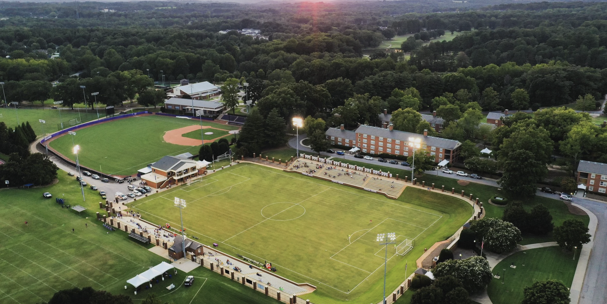 Aerial view of a sports complex with baseball and soccer fields, illuminated by stadium lights, surrounded by trees and some residential buildings.