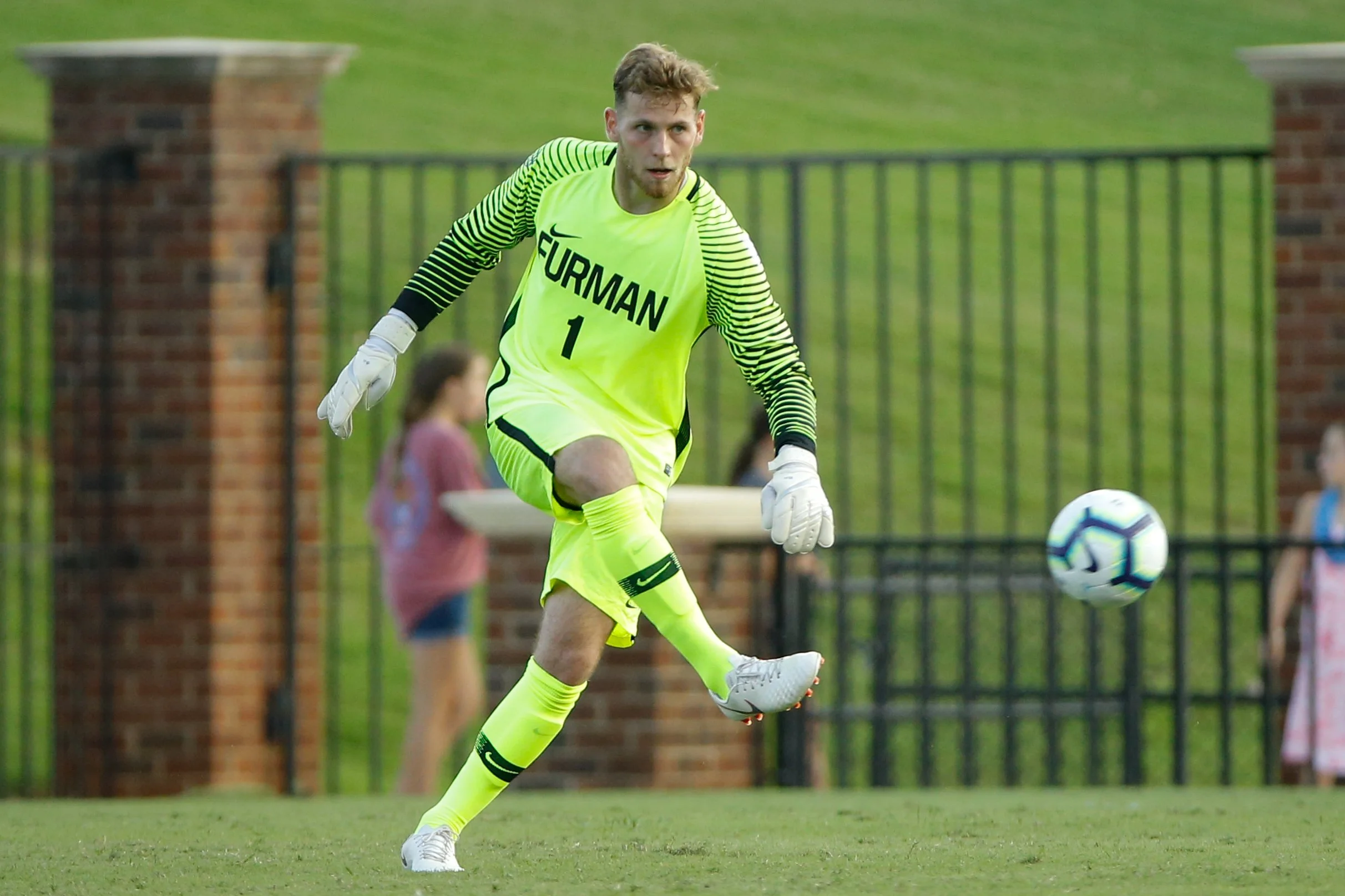 A male soccer goalkeeper in a bright yellow jersey with black lettering and stripes, wearing white gloves, is kicking a soccer ball on a green field with a black fence and brick pillars in the background.
