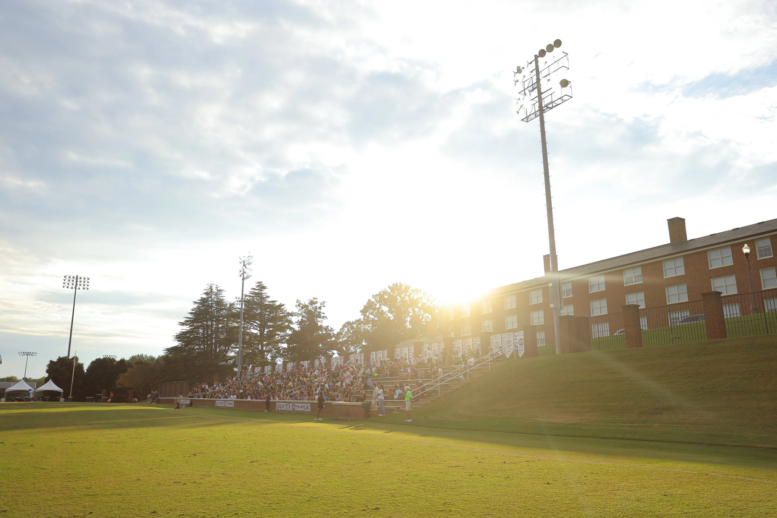 People sitting on bleachers at a sports field during sunset, with a building and tall floodlights in the background.
