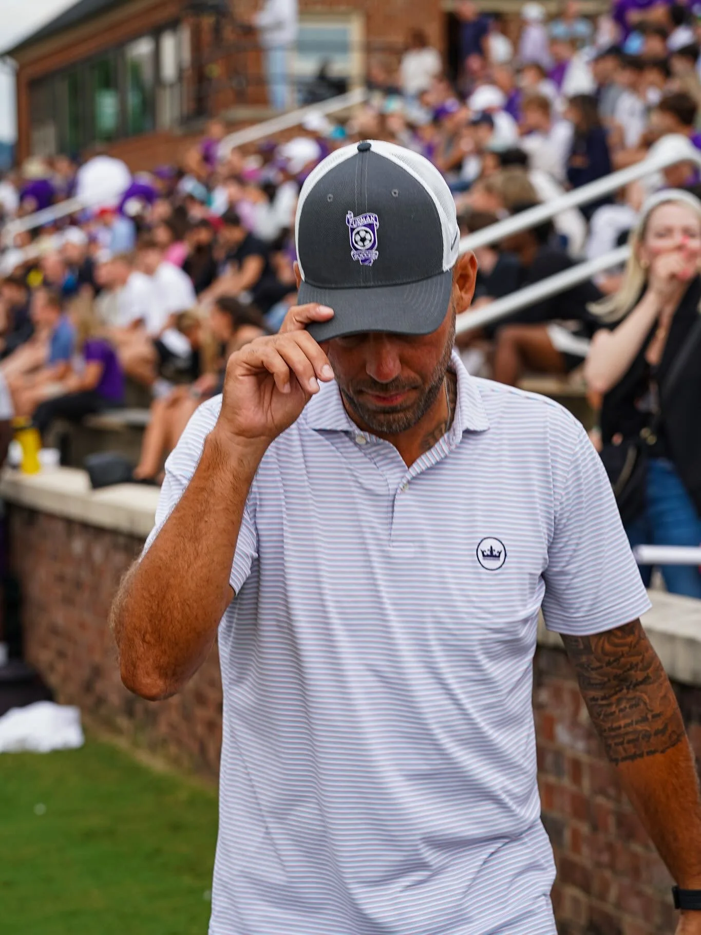 A man wearing a striped polo shirt and a gray cap is standing outdoors at a sports event, with a crowded stadium background.