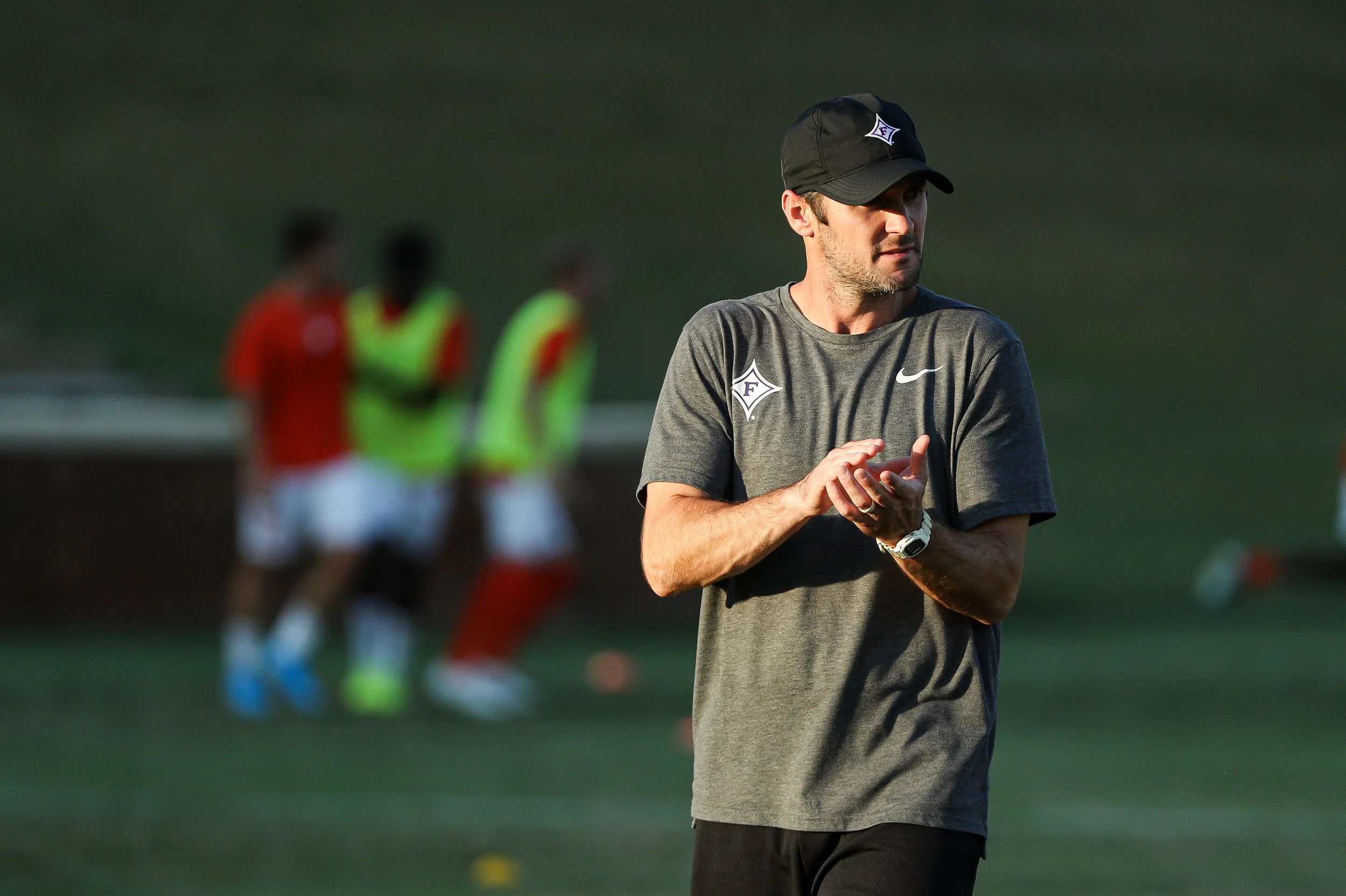 A man in a gray Nike t-shirt and black baseball cap, clapping his hands during a soccer practice on a grassy field with blurred players in the background.