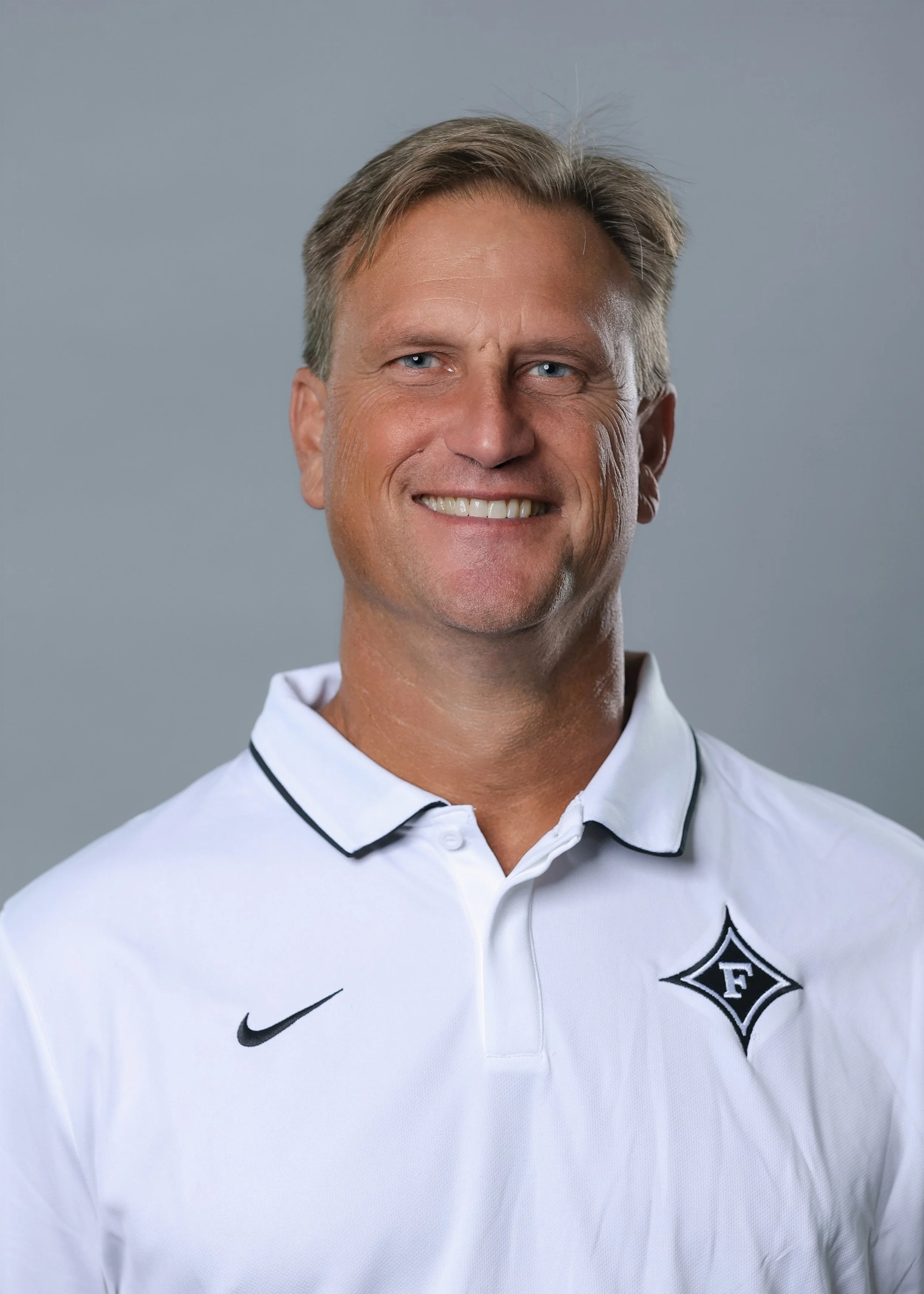 Portrait of a smiling middle-aged man with blond hair, wearing a white sports polo shirt with a black Nike logo and a black and white diamond-shaped emblem on the left side.