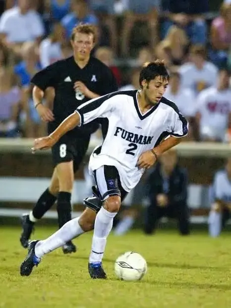 A soccer player from Furman University wearing a white jersey with black accents, number 2, dribbling a soccer ball on the field during a game. A referee and spectators are visible in the background.
