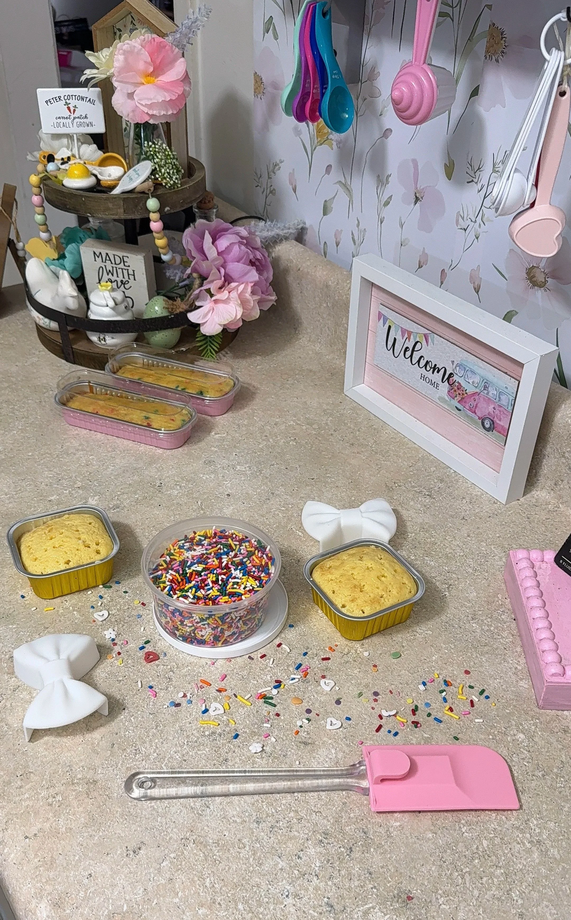 Decorative kitchen counter with colorful sprinkles, cupcake charms, containers of sprinkles and baked goods, pink and white bows, and a greeting sign that says 'Welcome Home'.