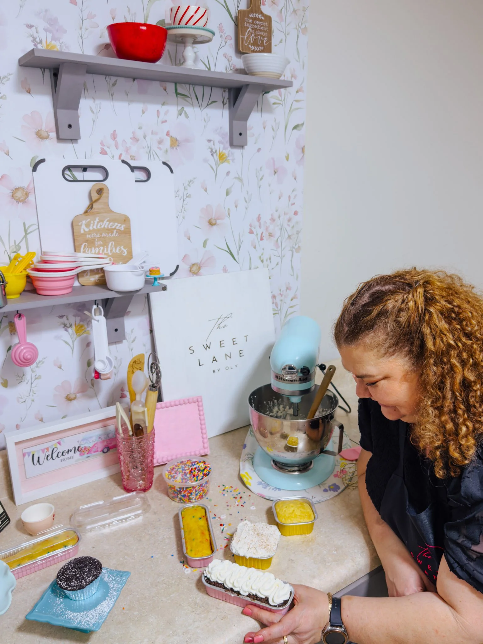 A woman in a bakery or kitchen decorating cakes with colorful sprinkles, surrounded by cake slices and baking supplies on a beige countertop, with a pastel blue KitchenAid mixer and floral wallpaper in the background.