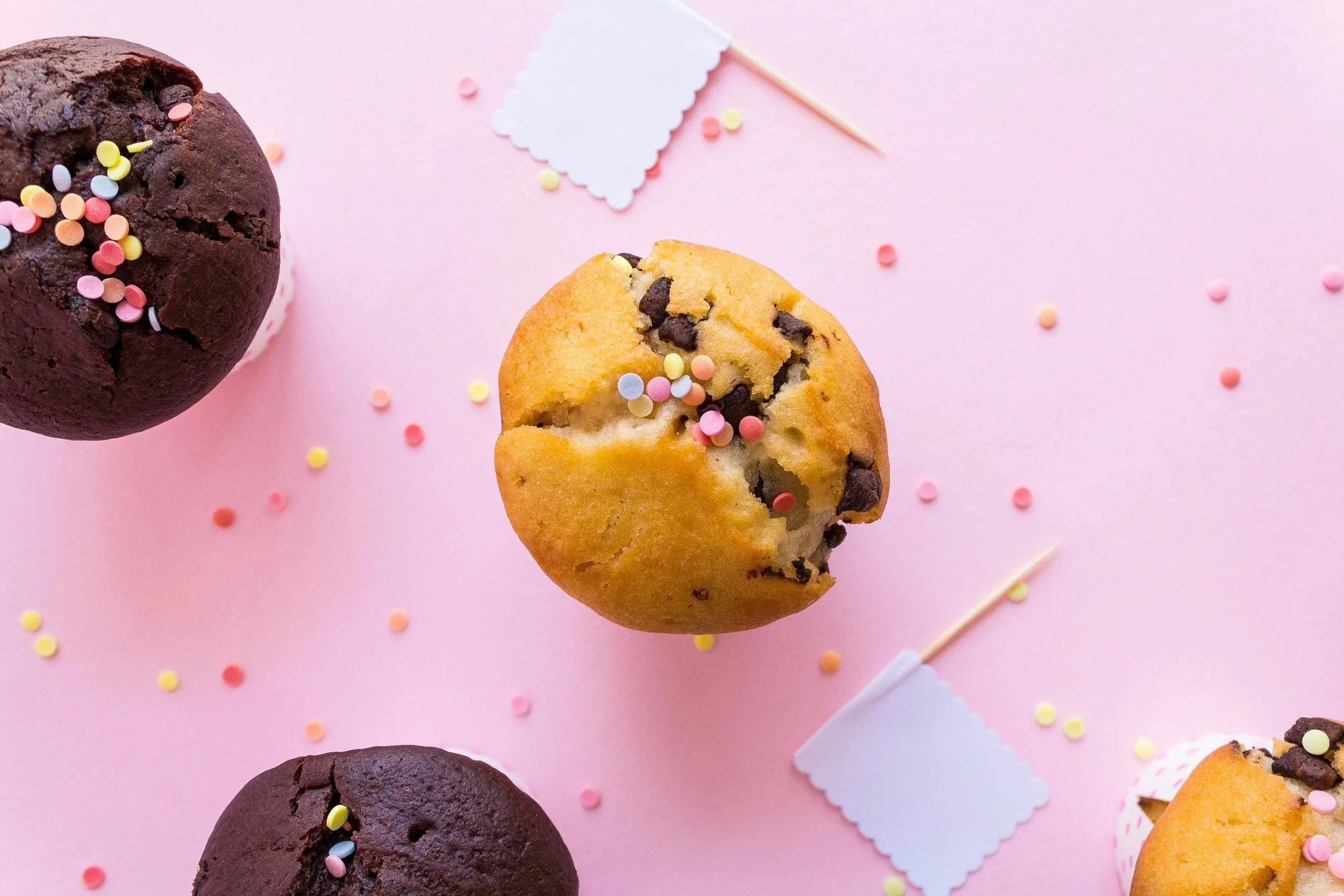 Chocolate chip muffins with colorful sprinkles on a pink background, some muffins in cupcake liners, and small blank flags scattered around.