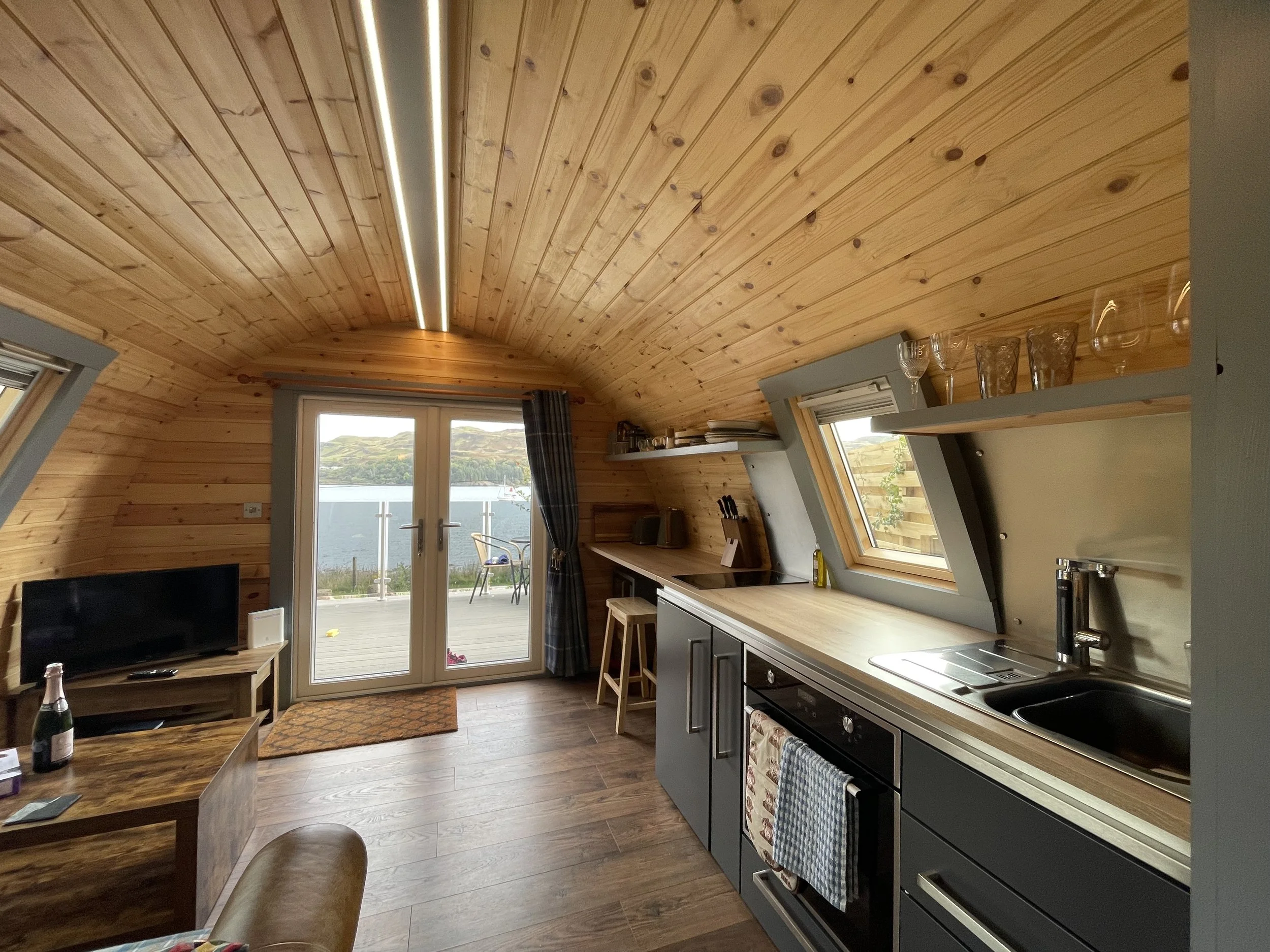 Interior of a cozy wooden cabin kitchen with a view of a lake outside, featuring a sliding glass door, a small TV, and a kitchen counter with cabinets, a sink, and windows above.