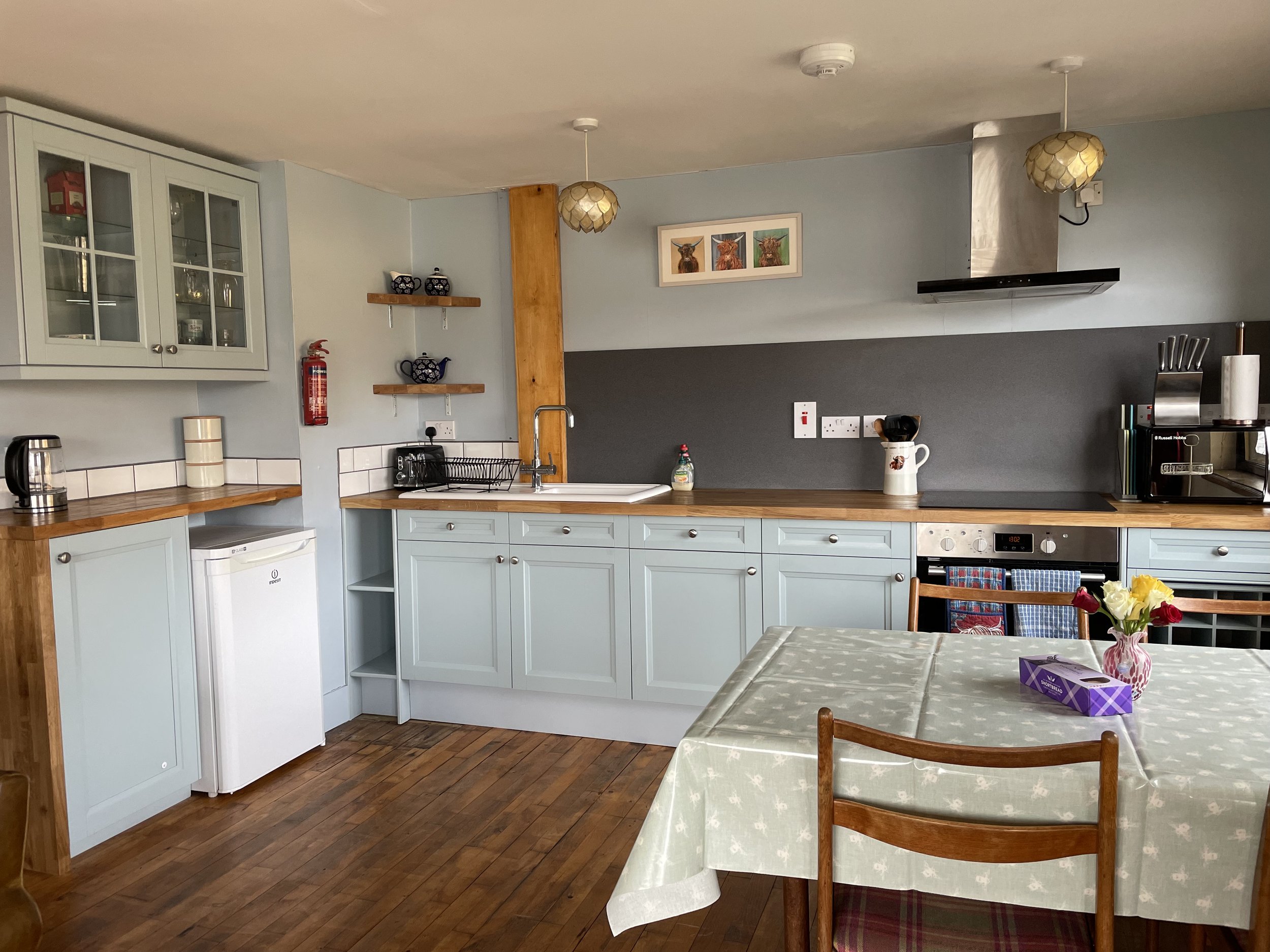 A kitchen with light blue cabinets, wooden countertops, a white mini fridge, a black toaster, and a dining table with a floral tablecloth and a vase of flowers.