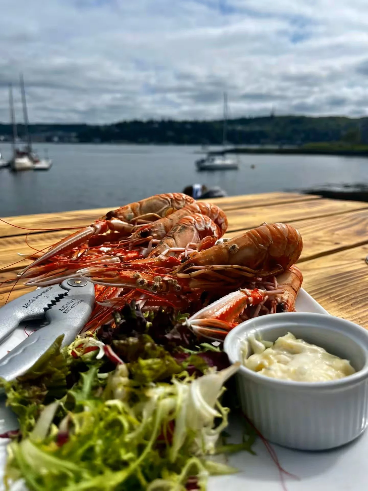 Plate of cooked lobster and shrimp with a side salad and dipping sauce, set on a wooden table outdoors near a body of water with boats and a cloudy sky.