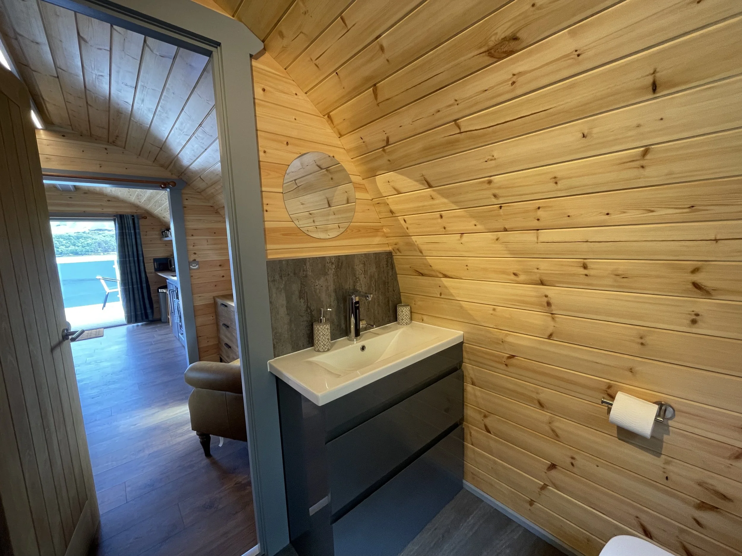 A small bathroom with wood-paneled walls and ceiling, a rectangular sink with gray drawers underneath, a mirror with an open circle cutout, and a toilet paper roll on the wall. The bathroom opens to a room with a balcony, curtains, a chair, and a view of water and trees outside.