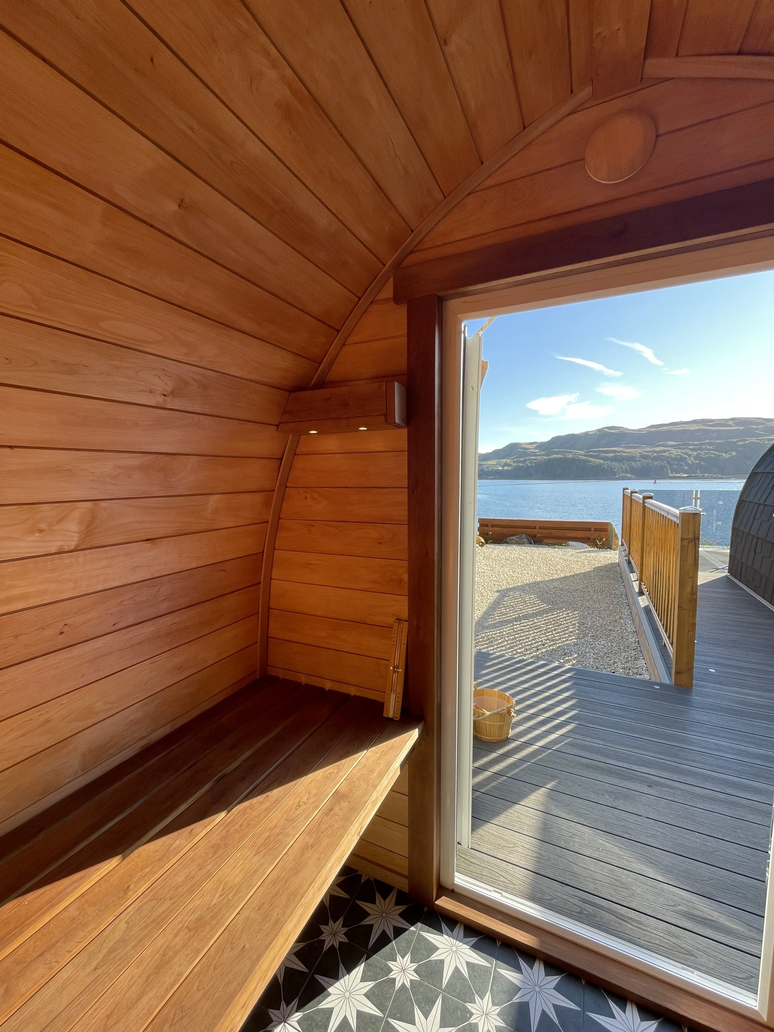 Interior of a wooden sauna with an open door leading to a deck overlooking water and mountains.