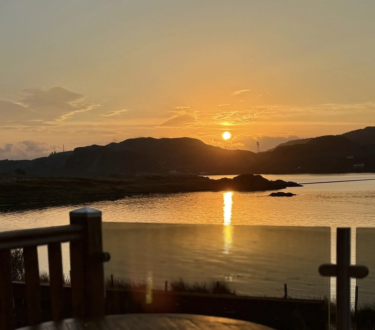 Sunset over a body of water with hills in the background, reflection of the sun on the water, and a wooden railing in the foreground.