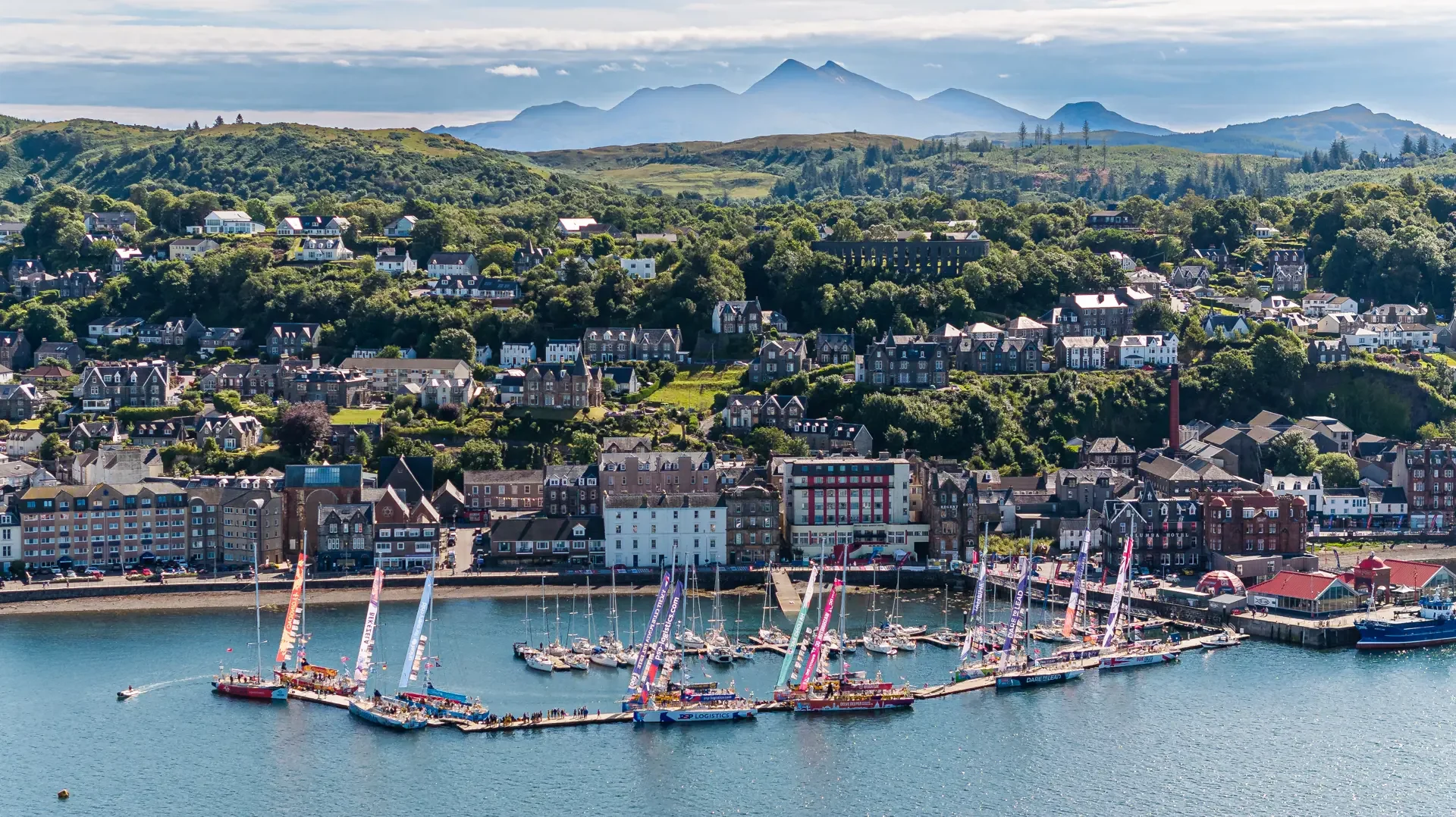A scenic coastal town with colorful sailboats docked at a marina, surrounded by a row of buildings, lush green hilly landscape, and mountains in the background.