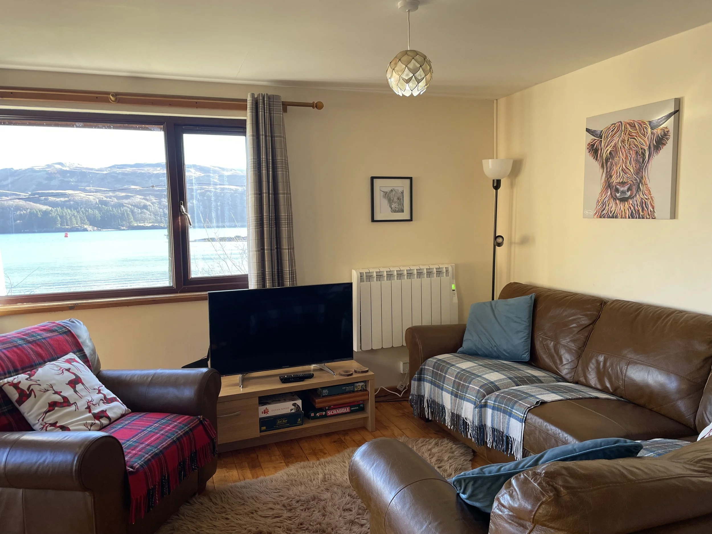 Living room with large window overlooking a lake and mountains, brown leather sofas, wooden TV stand with TV, picture frames on the wall, floor lamp, beige walls, and a fluffy rug.