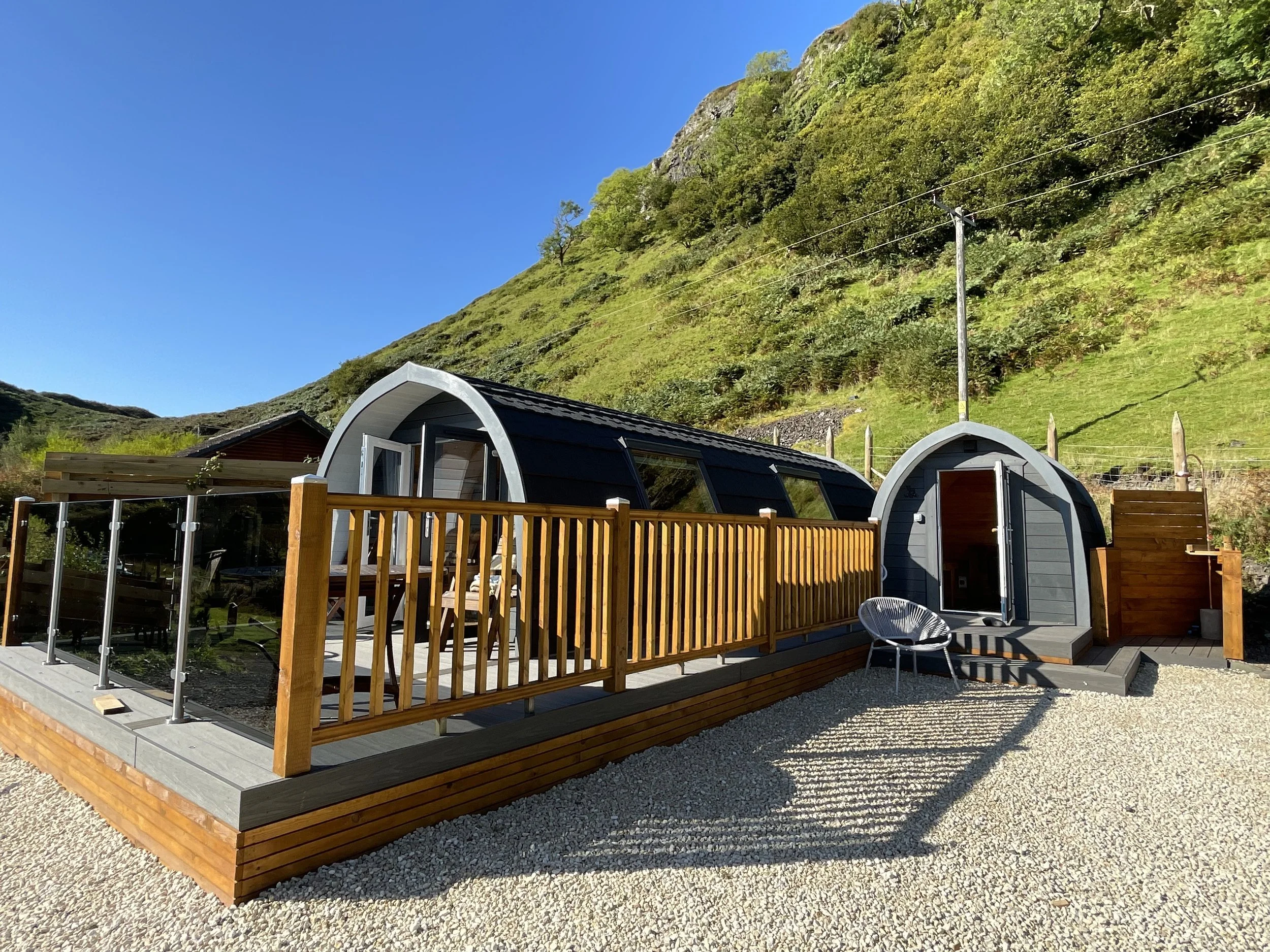 Photo of two small, modern, gray wood-clad cabins with arched roofs, situated on a gravel surface at the base of a lush green hillside under a clear blue sky.