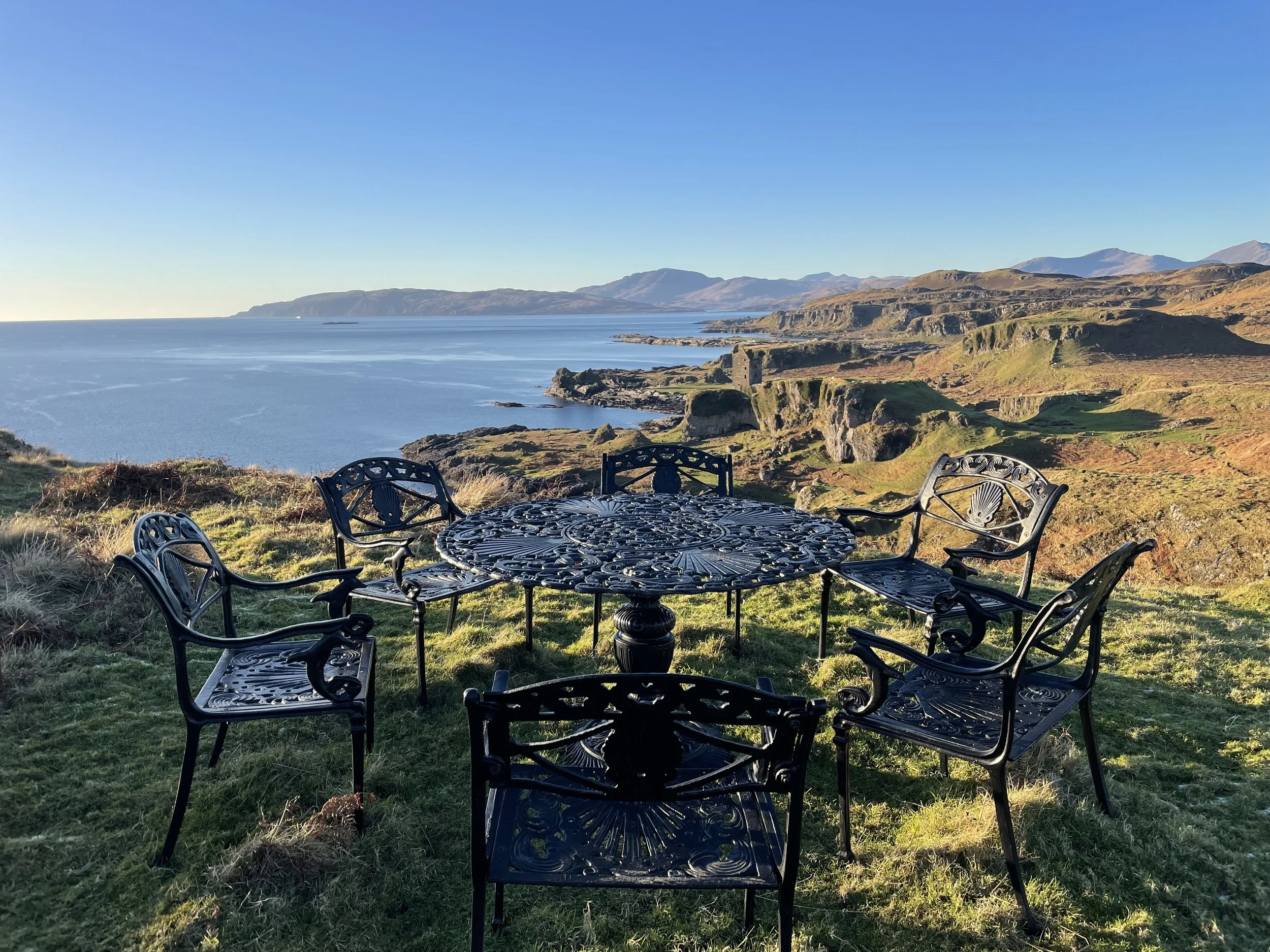 Outdoor cast iron table and five matching chairs on grassy hill with ocean and rugged coastline in background under clear sky.