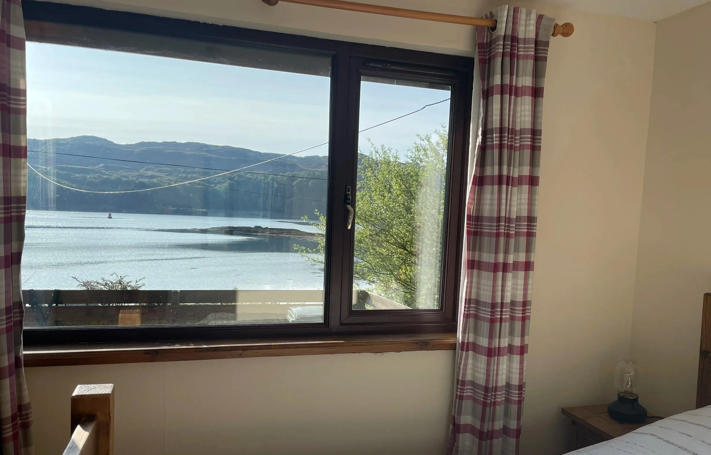View of a lake or sea through a window with hills in the background, outside a room with plaid curtains and a bedside table with a lamp.