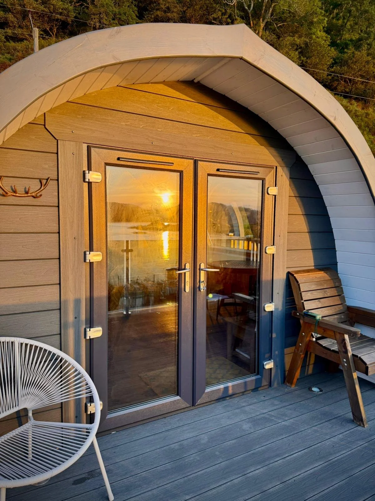 View of a wooden cottage with glass double doors reflecting a sunset over a river, with outdoor chairs on a wooden deck.