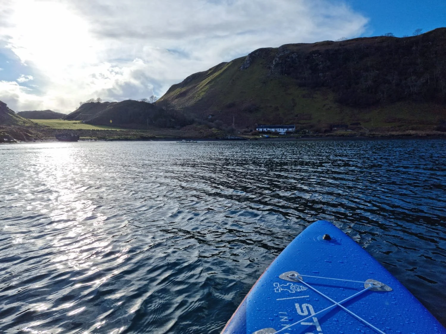 View from a paddleboard on a body of water, with green hills and mountains in the background under a partly cloudy sky.