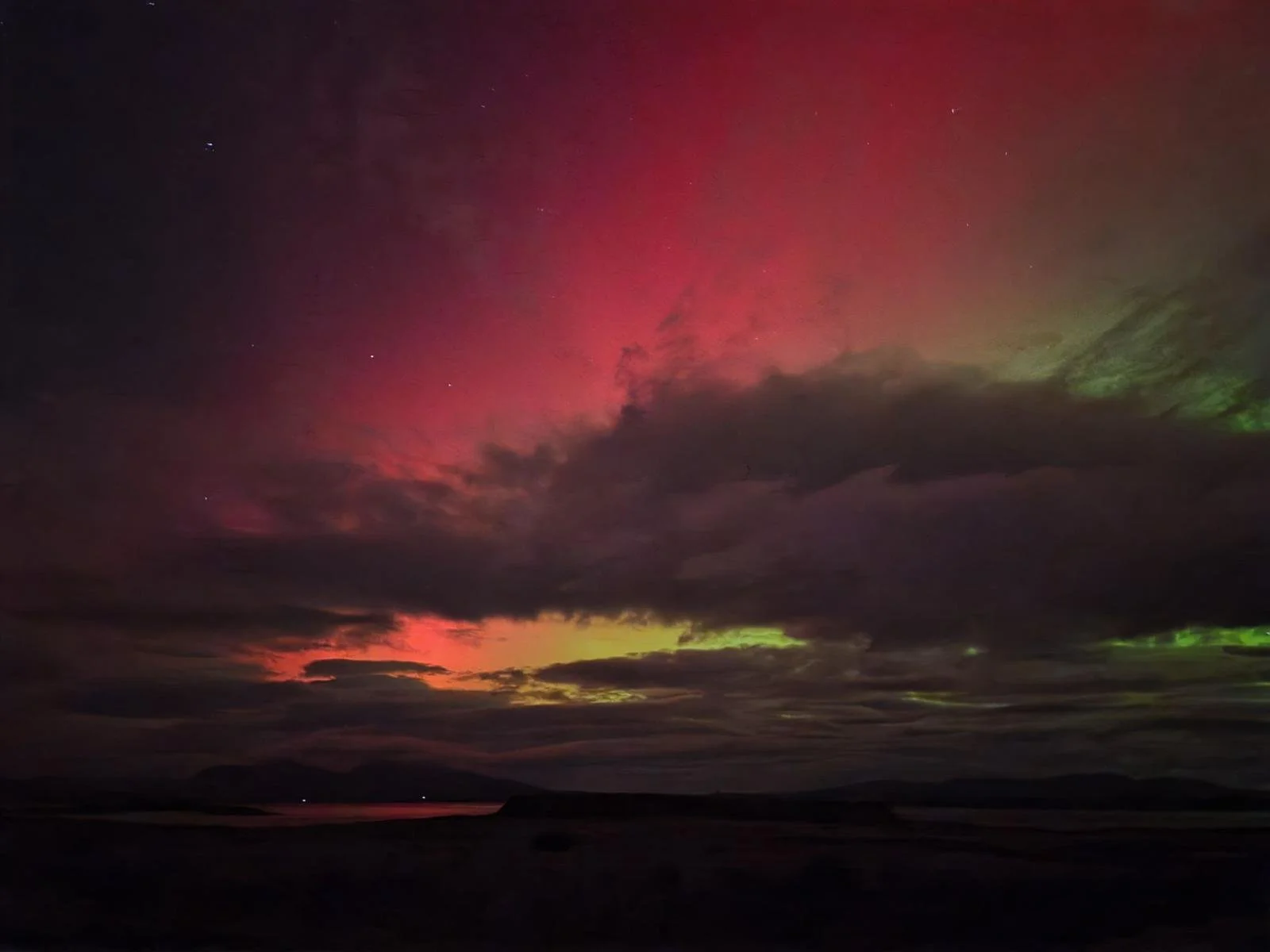 Night sky with colorful aurora borealis over a dark landscape with water and hills.