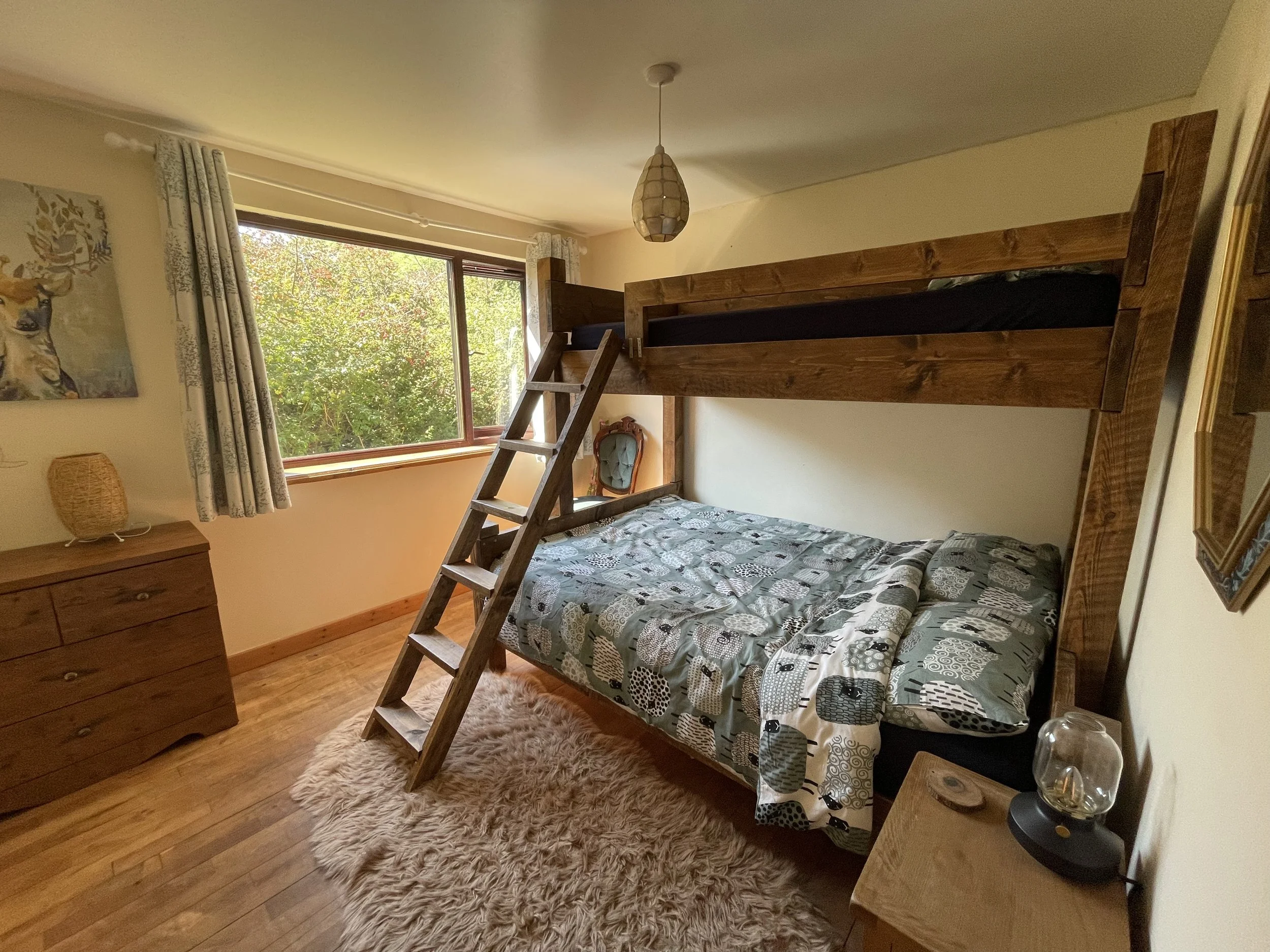 Bedroom with wooden bunk bed, window with green foliage outside, floral curtains, wooden dresser, small bedside table with lamp, and artwork on the walls.