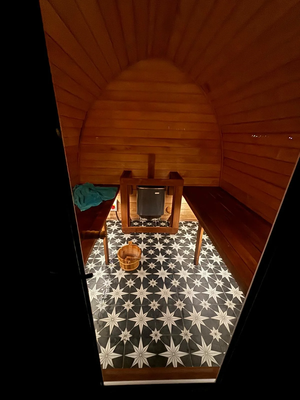 A small, wooden sauna room with a curved ceiling. It has two wooden benches on either side, a heater in the center, and a bucket on the patterned floor. There is some clothing on the left bench.