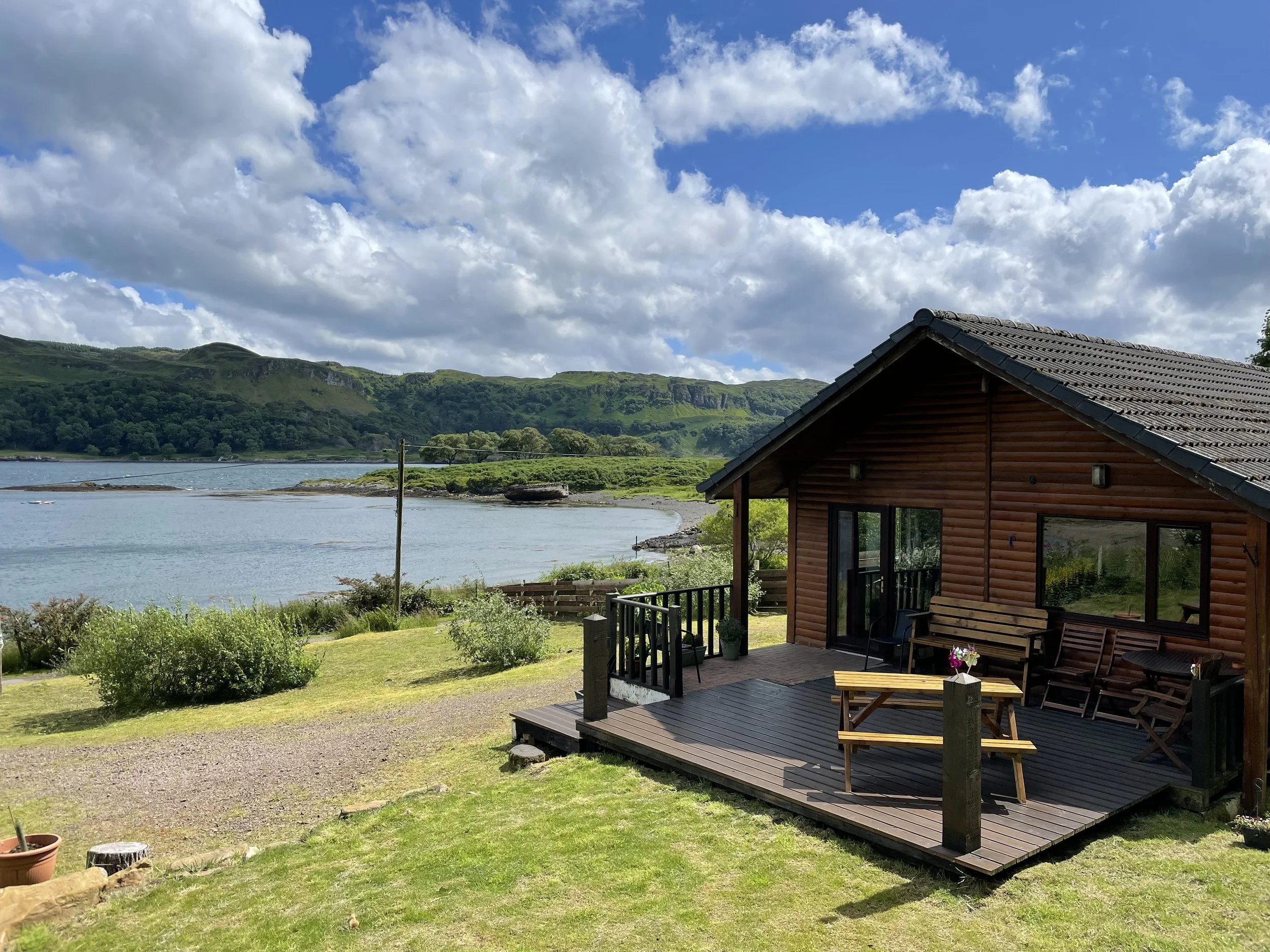 A wooden cabin with a deck overlooking a lake, surrounded by green hills and a partly cloudy sky.
