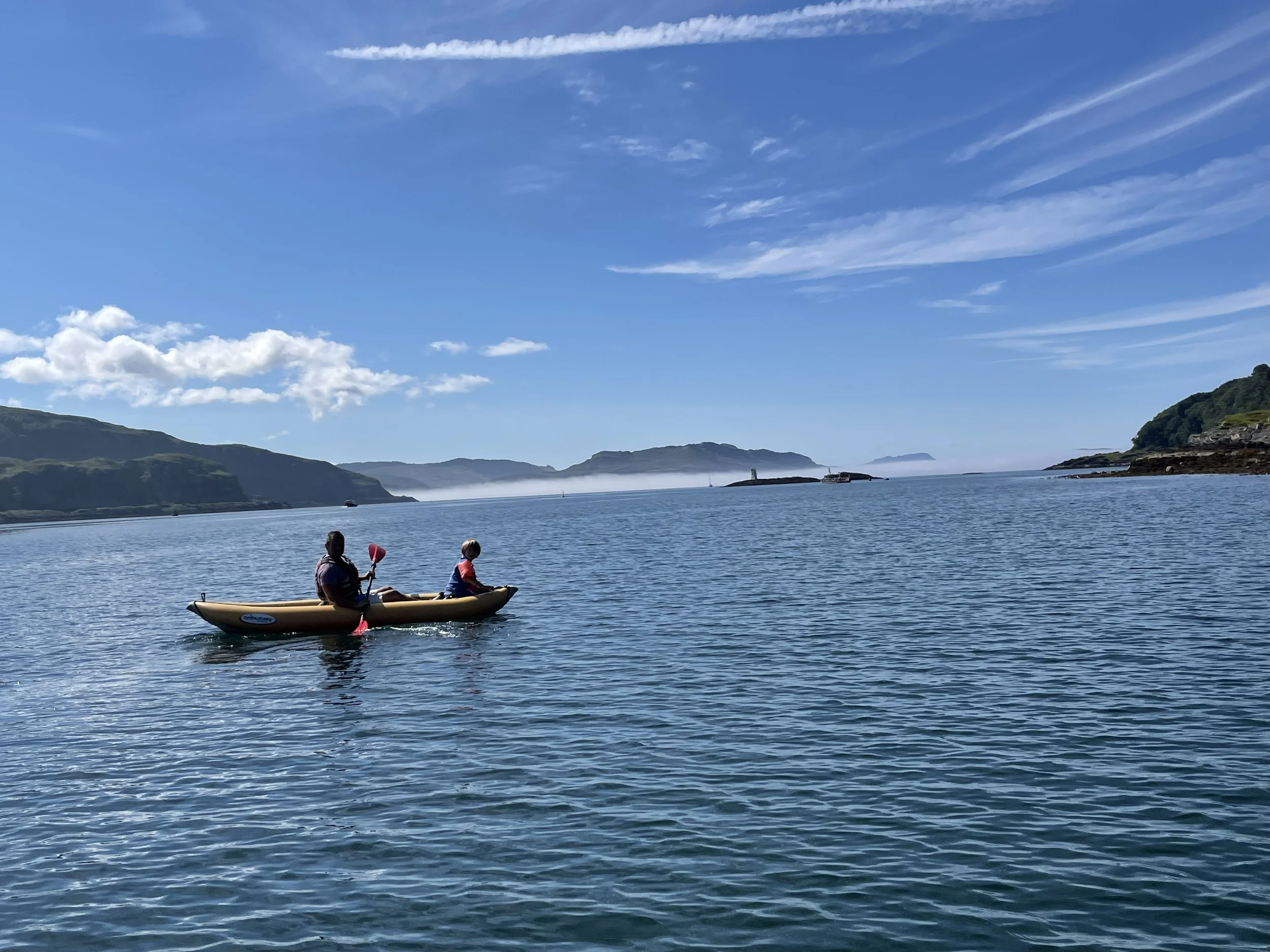 Two children kayaking on calm water with a scenic coastline and hills in the background, under a blue sky with wispy clouds.