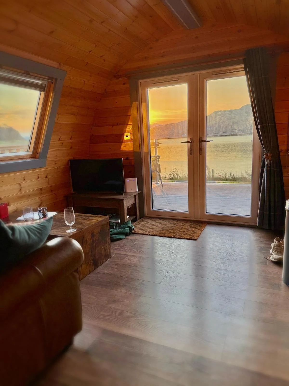 Cozy wooden interior of a room with a view of a sunset over water and mountains through glass doors, with a brown leather sofa, a small wooden table, a TV, and a sliding door with curtains.