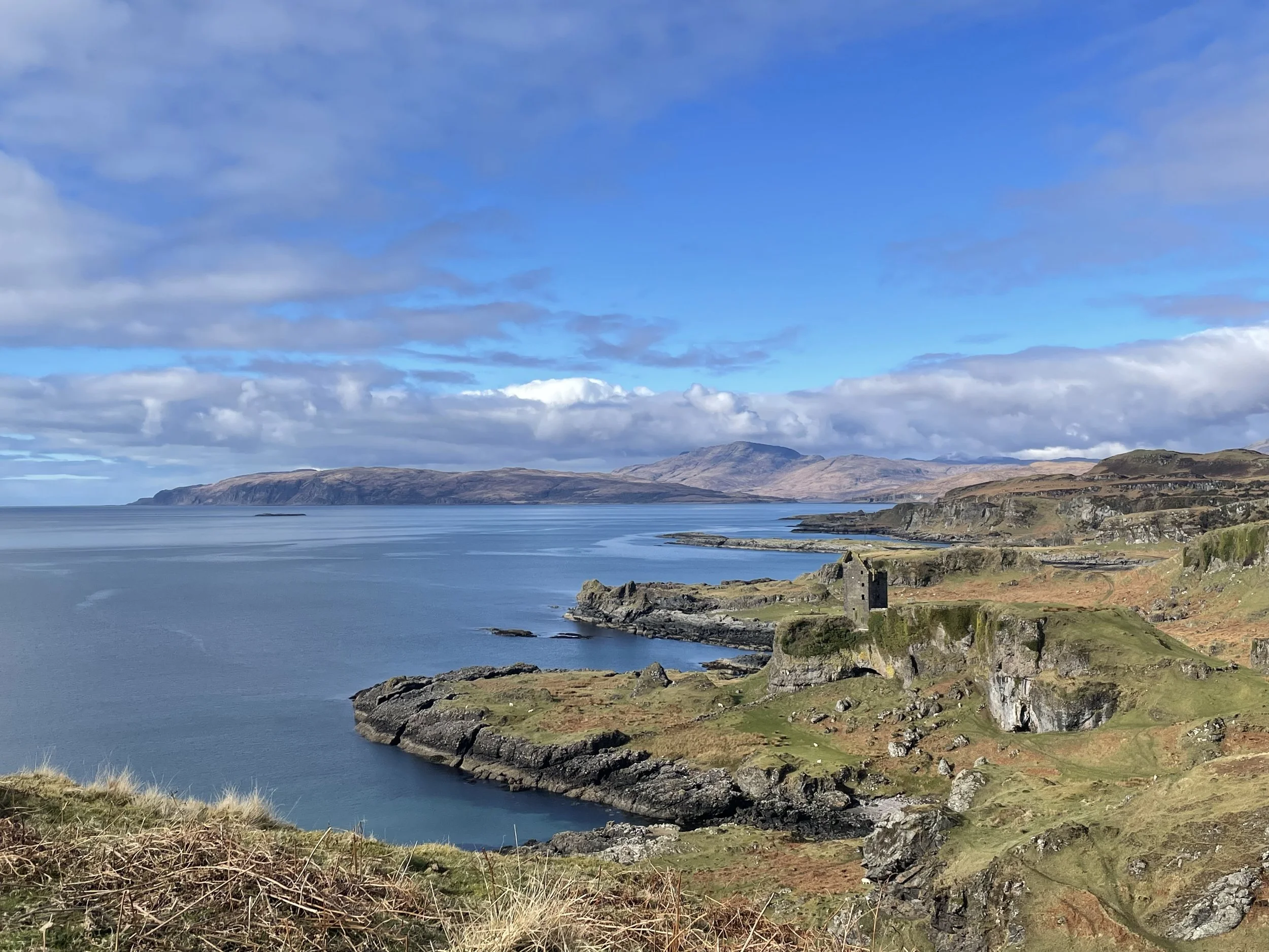 Scenic coastal landscape with rugged cliffs, a small stone building, overlooking a calm blue sea under a partly cloudy sky, with distant mountains in the background.