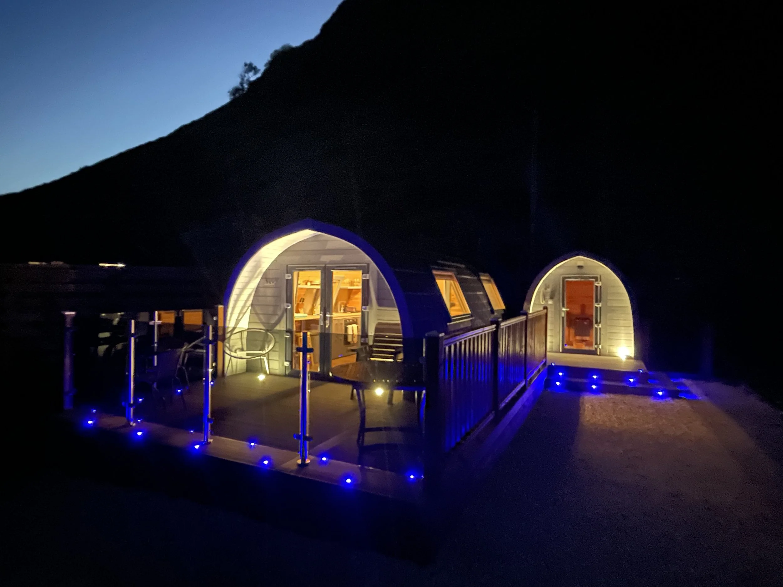 A small house with a rounded shape, illuminated from inside, with blue LED lights along the deck railing and steps, set against a dark mountain landscape at night.