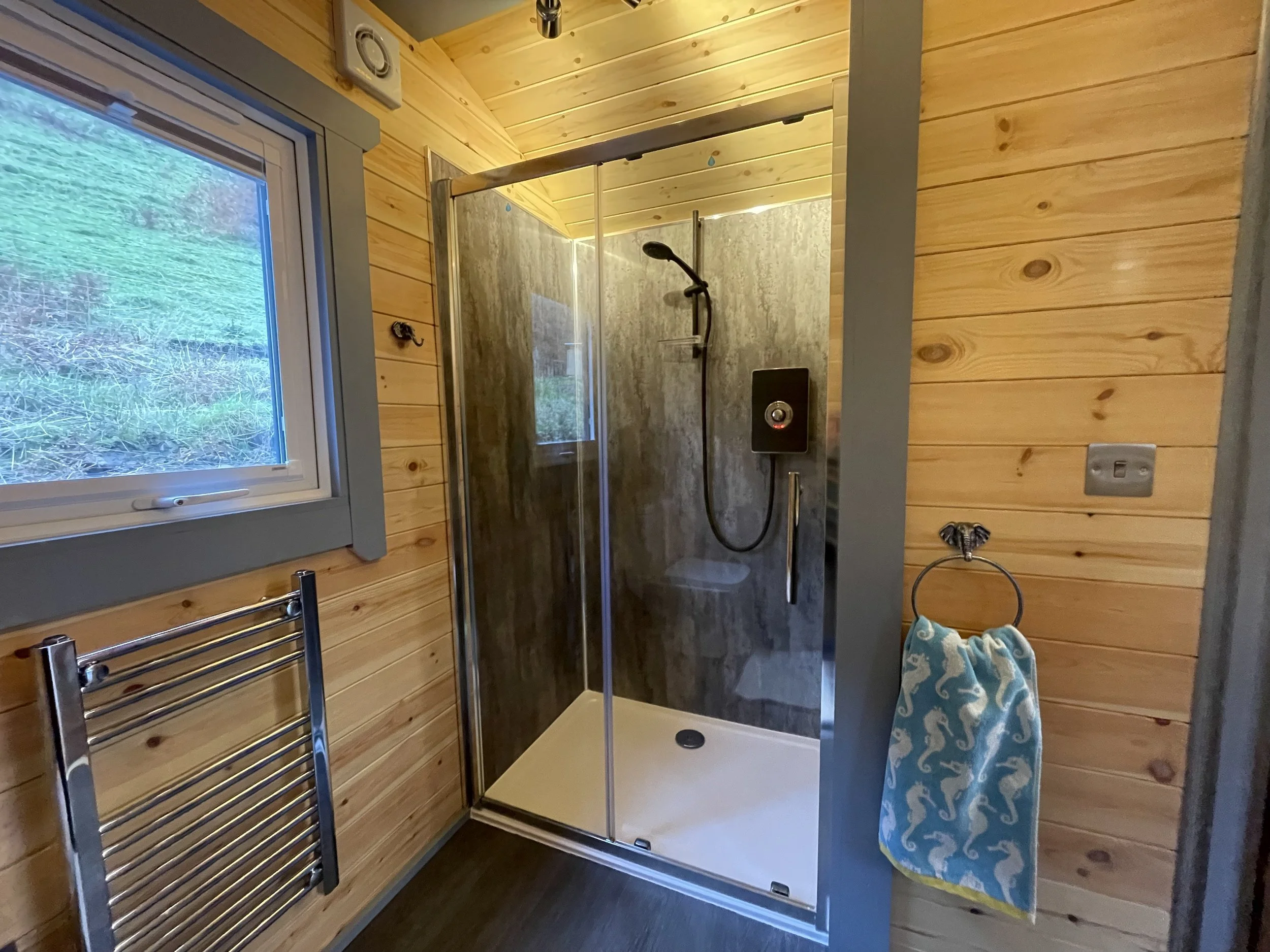 Small shower area with a water-resistant steamy wall, black showerhead, a small window to the left with a view of outside greenery, wooden plank walls, a towel with sea creatures hanging on a hook, and a chrome towel rack below the window.