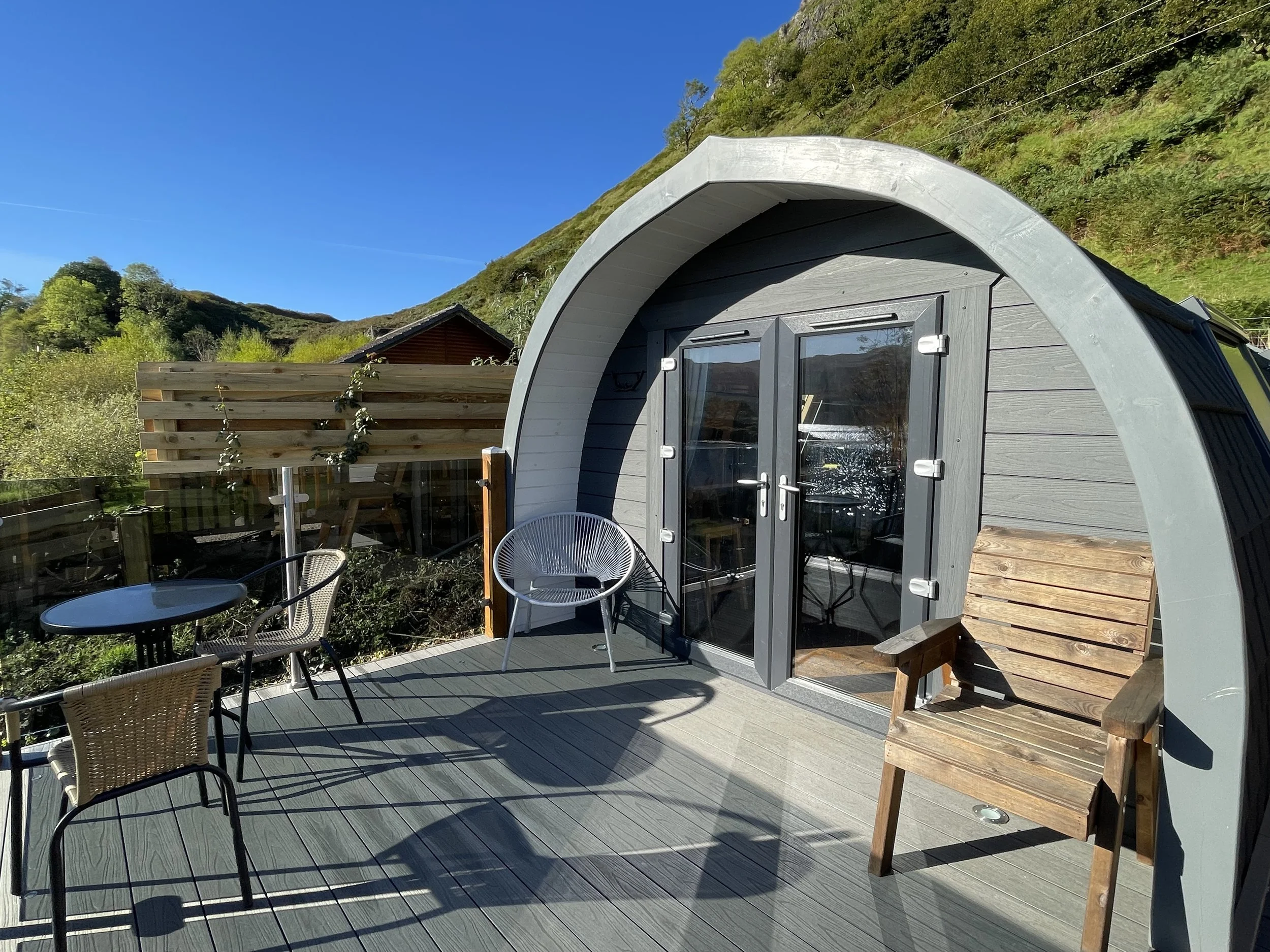 Outdoor balcony with chairs and a wooden cabin, scenic hillside in the background, bright and sunny weather.