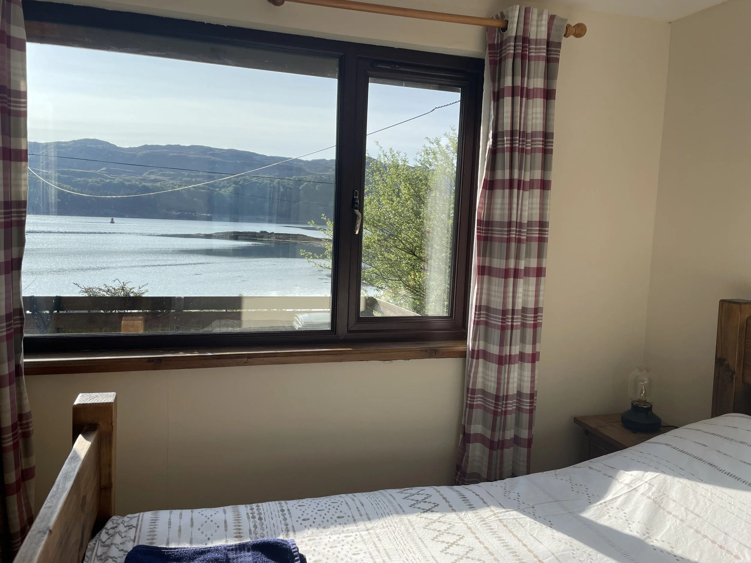 A bedroom with a large window looking out onto a lake and mountains, curtains with a red and white checkered pattern, a wooden bed frame, a white patterned bedspread, and a bedside table with a glass lamp.
