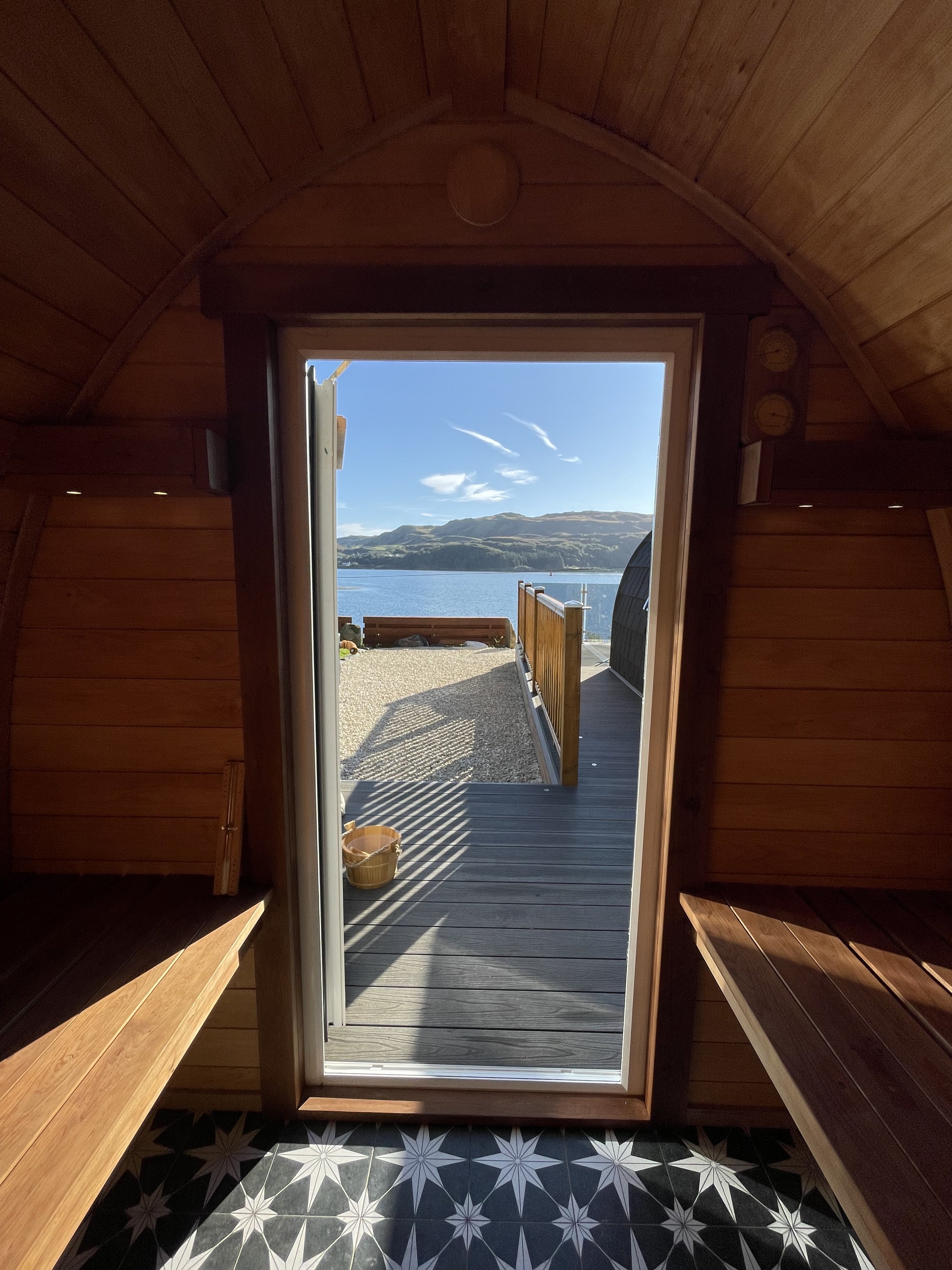 View from inside a wooden sauna looking out through an open door to a deck with a bench, overlooking a body of water and hills under a blue sky.