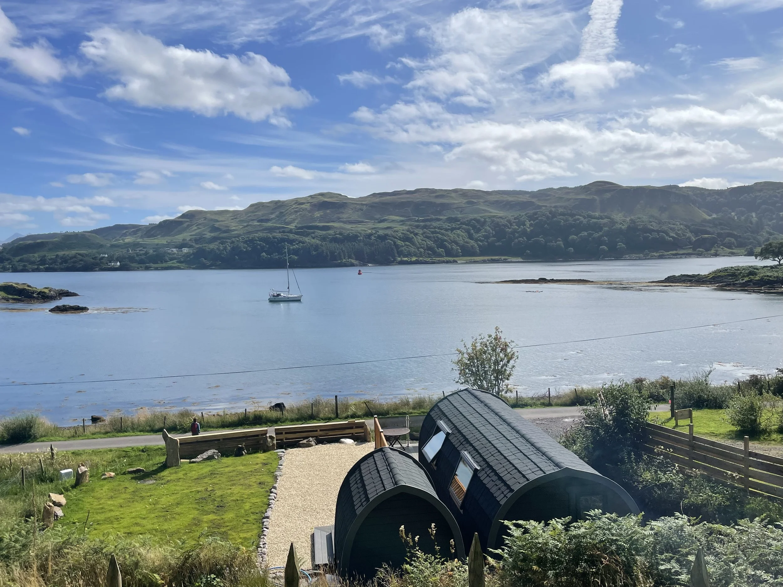 A peaceful lakeside scene with blue water, a boat, green hills in the background, and small black structures in the foreground surrounded by grass and a gravel pathway.