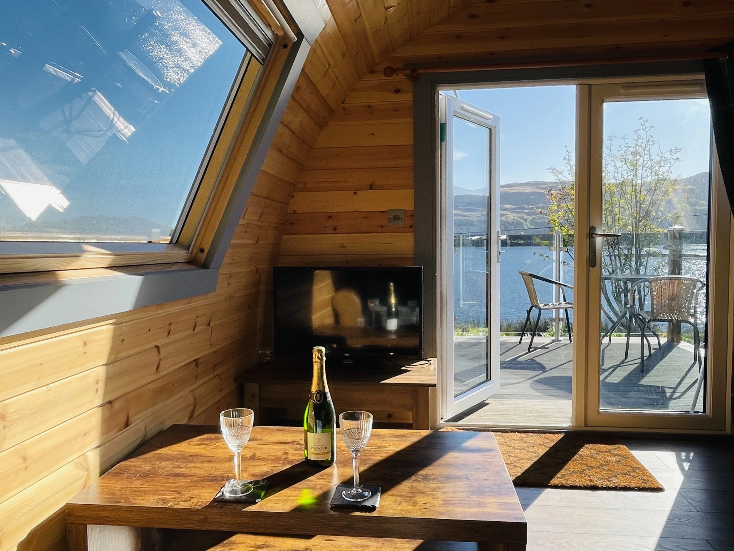 Cozy room with wooden walls, a small wooden table set with two empty glasses and a bottle of sparkling wine, a TV, and an open door leading to a balcony overlooking water and mountains on a sunny day.