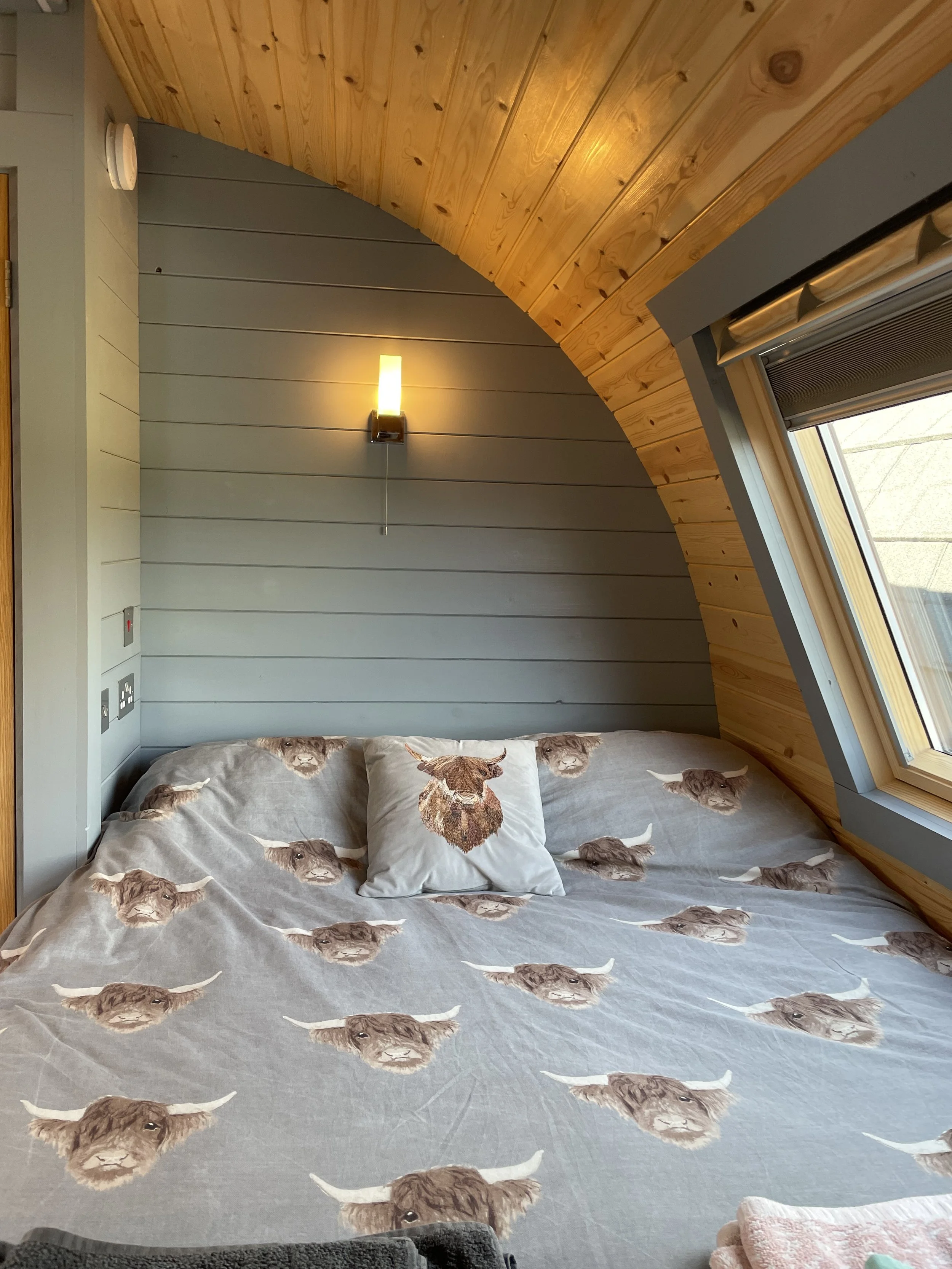 A cozy bedroom with a bed covered in a Highland cow-themed bedspread and matching pillow, set against a paneled wall with a wall-mounted light, and a large window with a sloped ceiling.