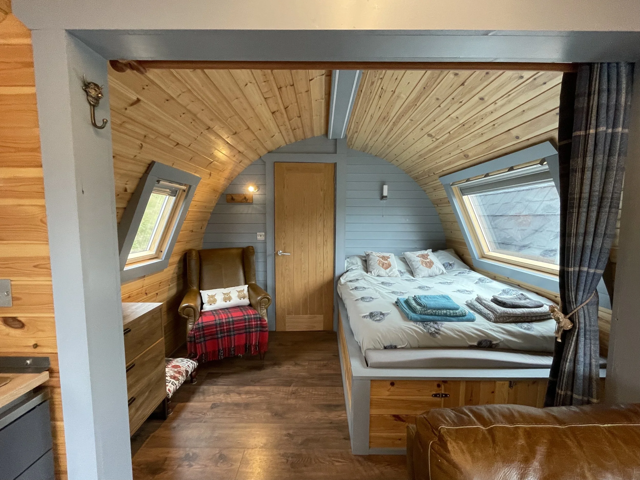 Cozy bedroom with sloped wooden ceiling, two skylight windows, a bed with woodland animal-themed bedding, an armchair with a red plaid blanket, and wood-paneled walls.