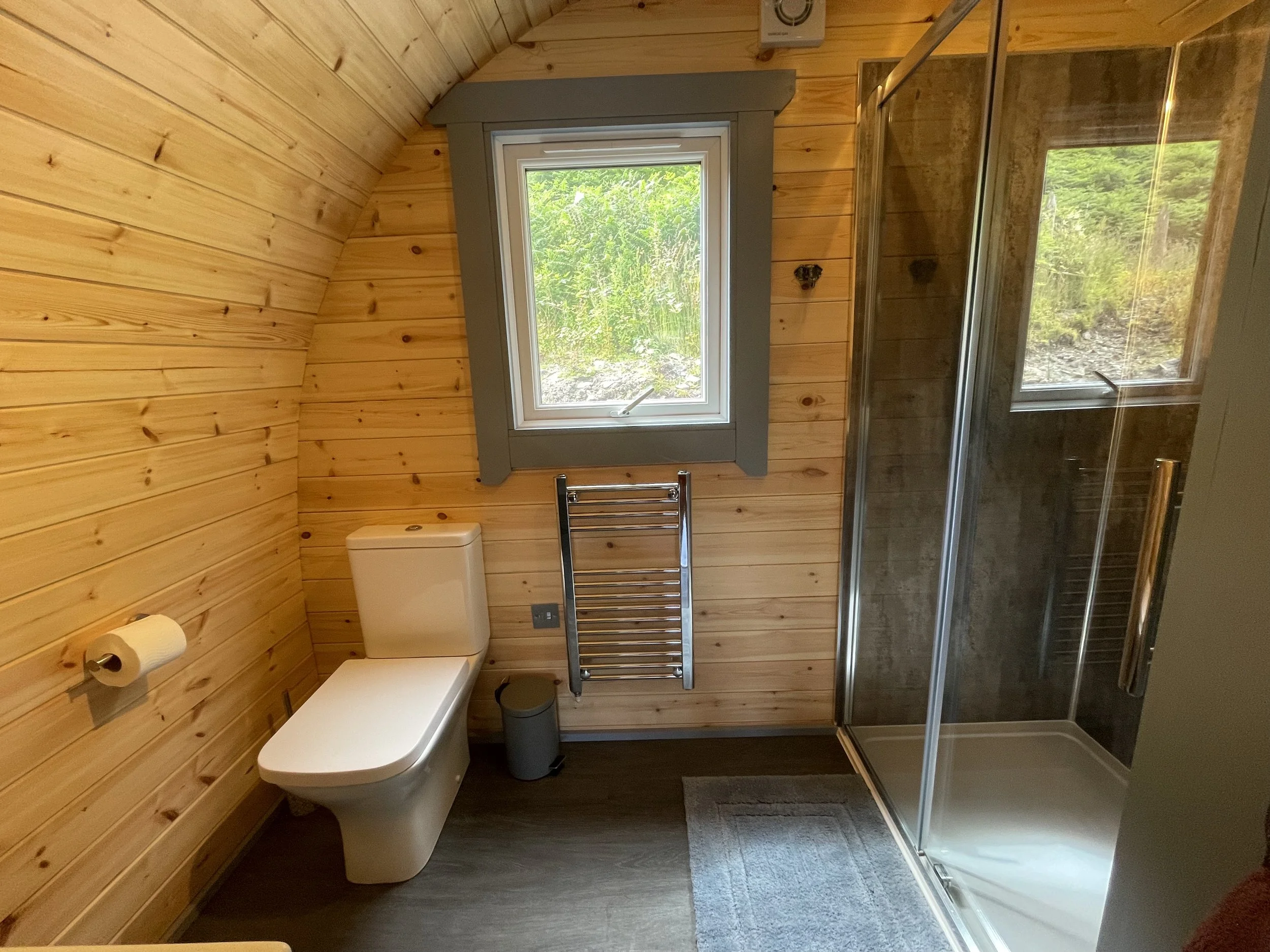 Wood-paneled bathroom with a toilet, a small trash bin, a heated towel rail, a window showing greenery, and a glass shower enclosure with a door handle.
