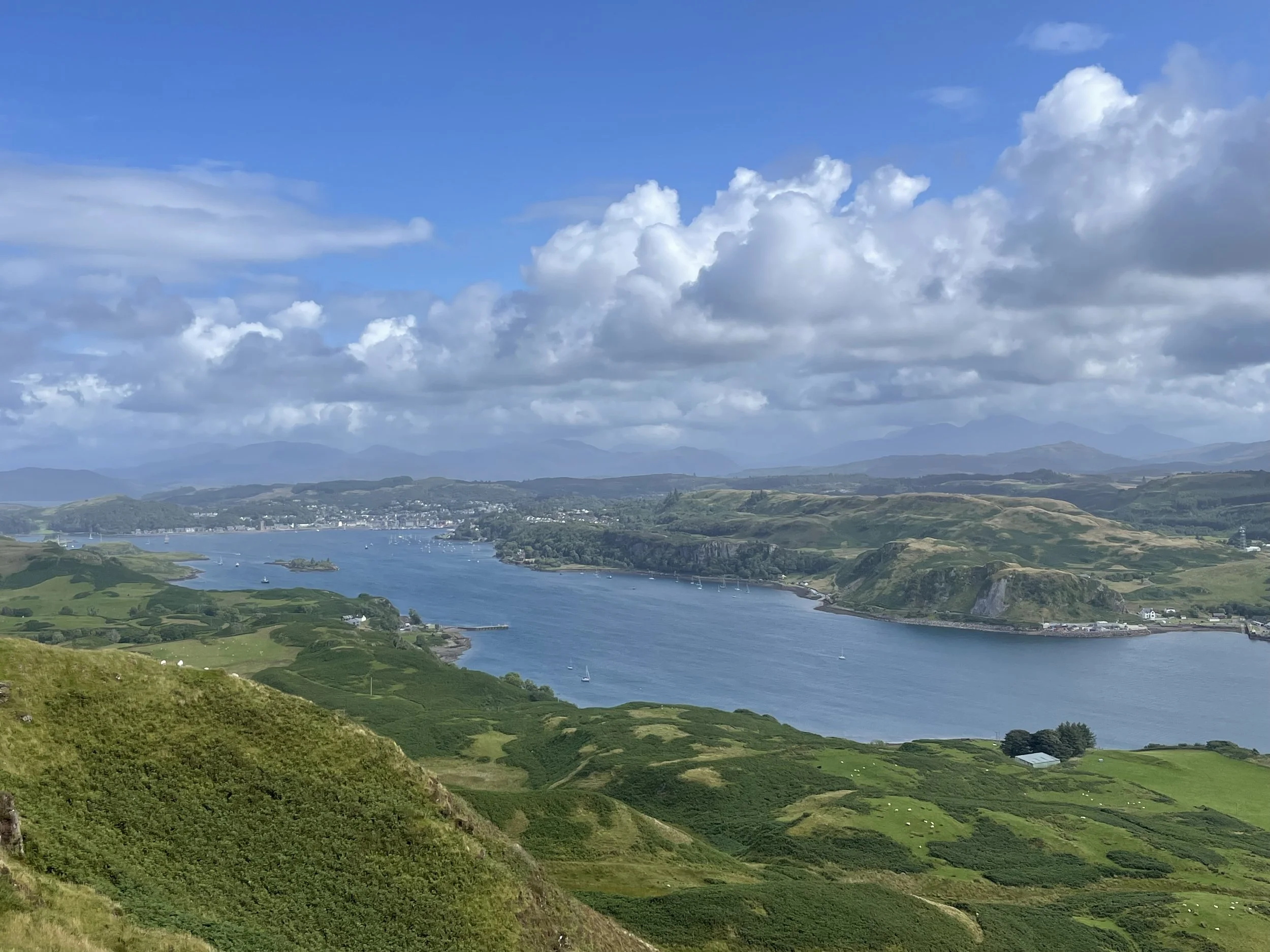 Scenic view of a river flowing through green hills and countryside with boats sailing, under a partly cloudy blue sky.
