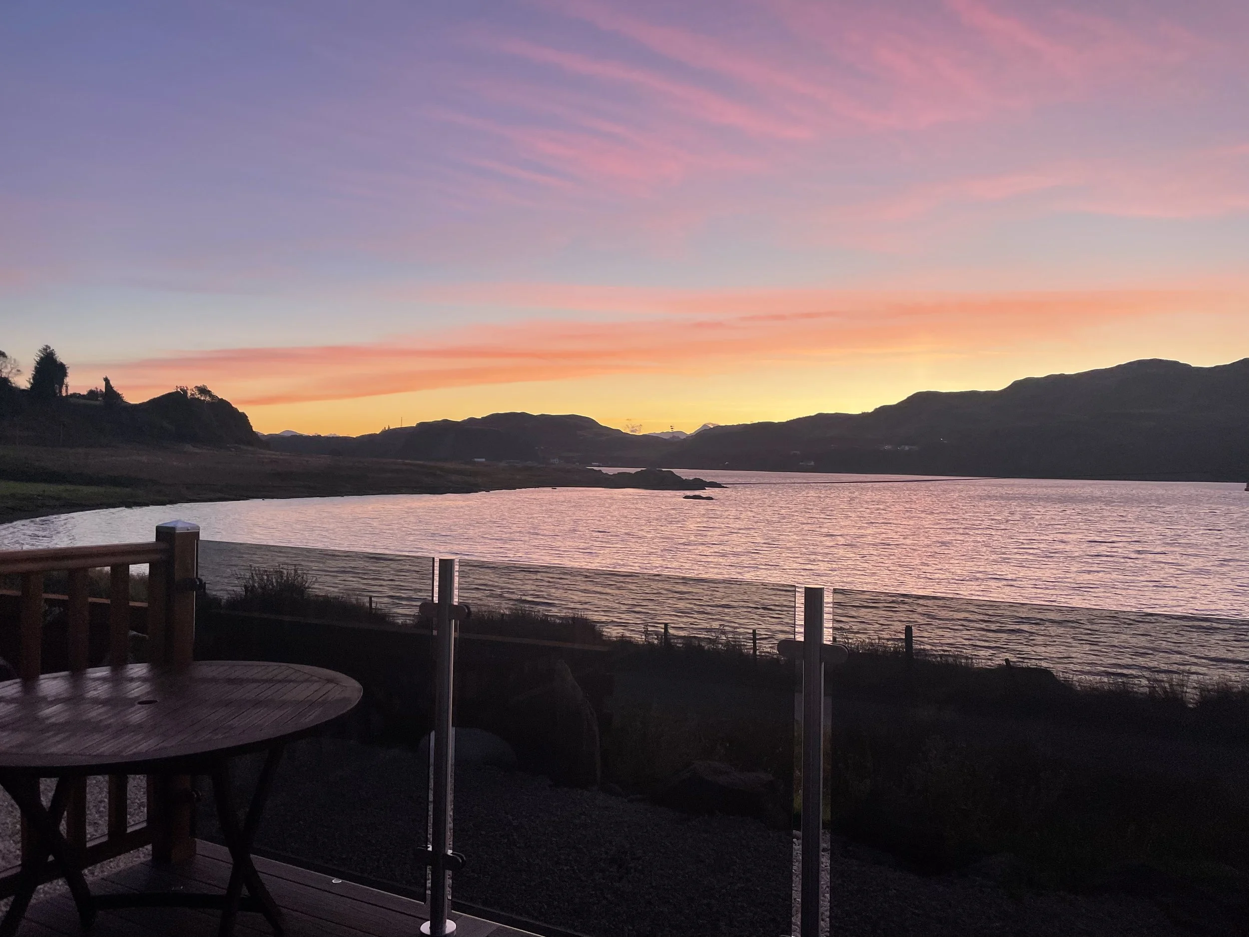 Sunset over a lake with pink, purple, and orange sky, mountains in the distance, and a wooden patio with a glass railing in the foreground.