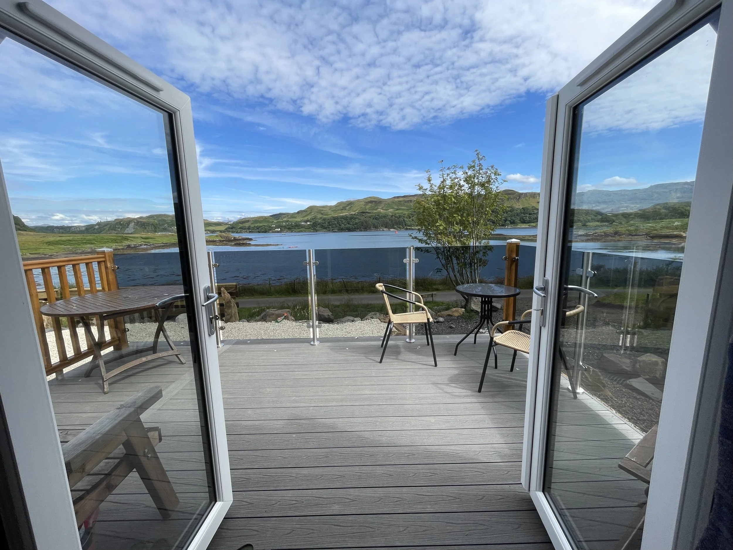 View of a wooden deck overlooking a lake with mountains in the distance, visible through open glass doors, with chairs and a small table on the deck, and a clear blue sky with some clouds.