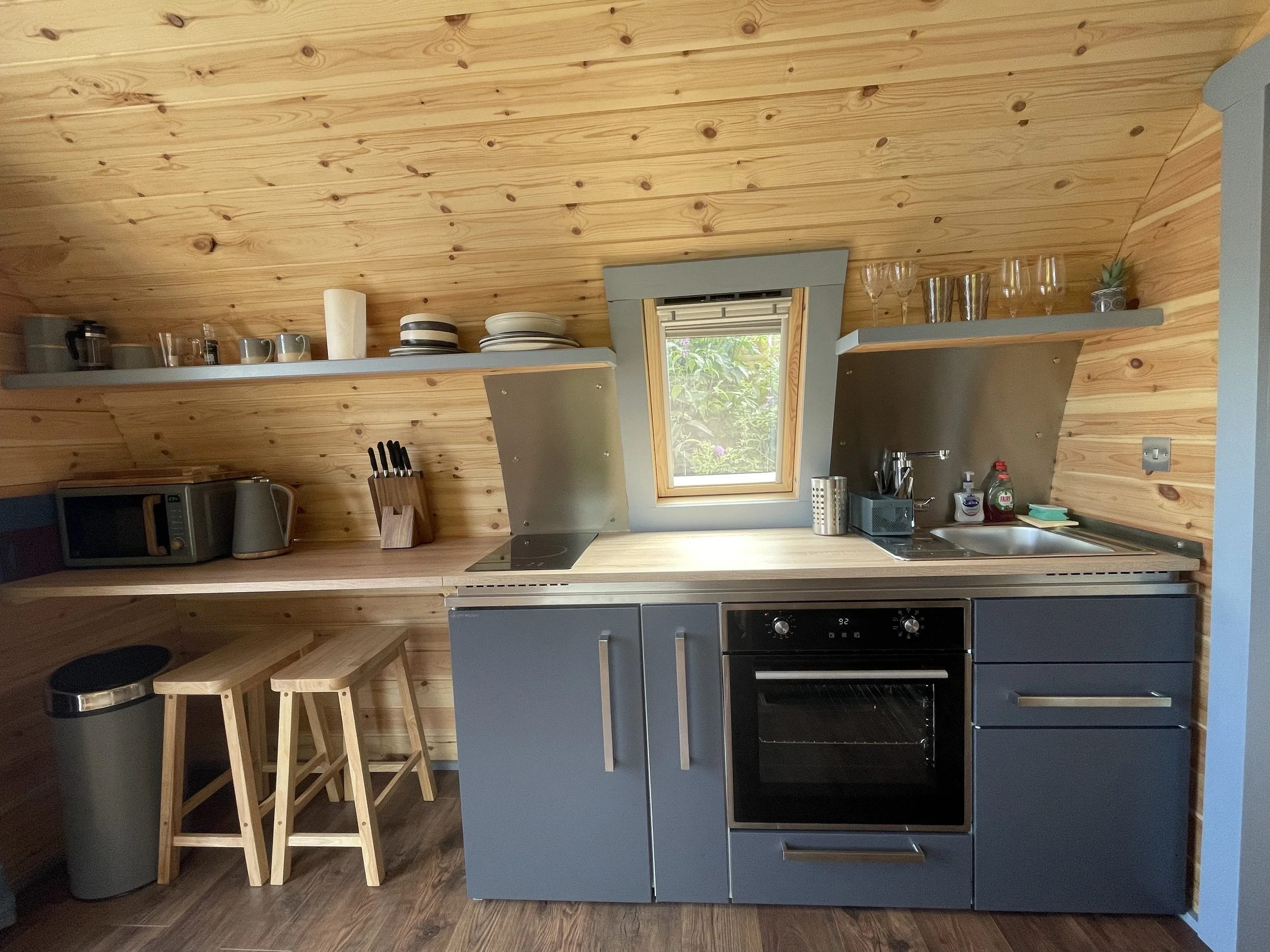 Small kitchen with wooden wall and ceiling, blue cabinets, black oven, stainless steel sink, window, shelves with dishes and glasses, microwave, kettle, kitchen tools, and stools.