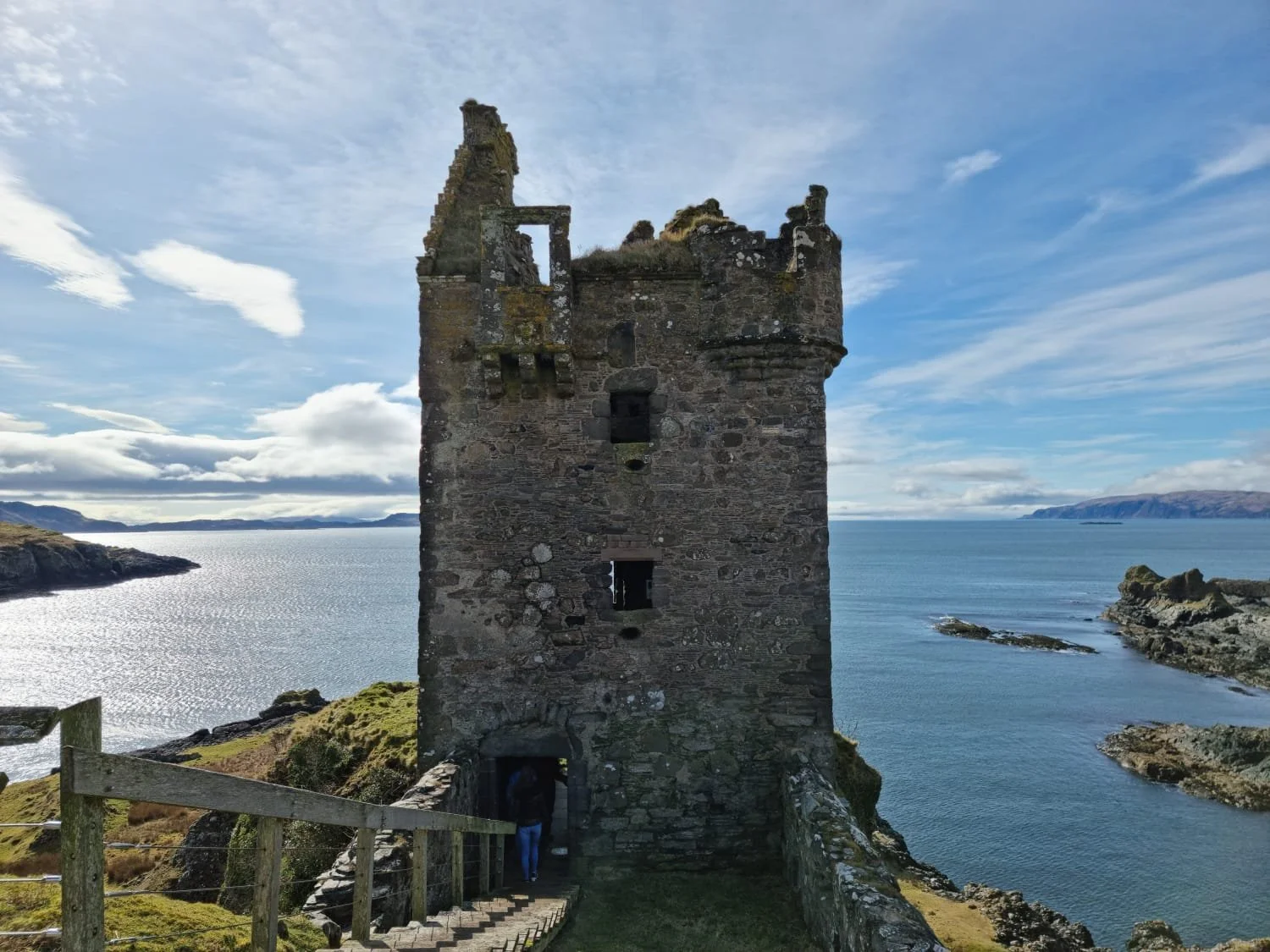 A tall, old stone tower on a hill overlooking the ocean, with a wooden walkway leading to its entrance. The sky is partly cloudy with sunlight reflecting off the water.