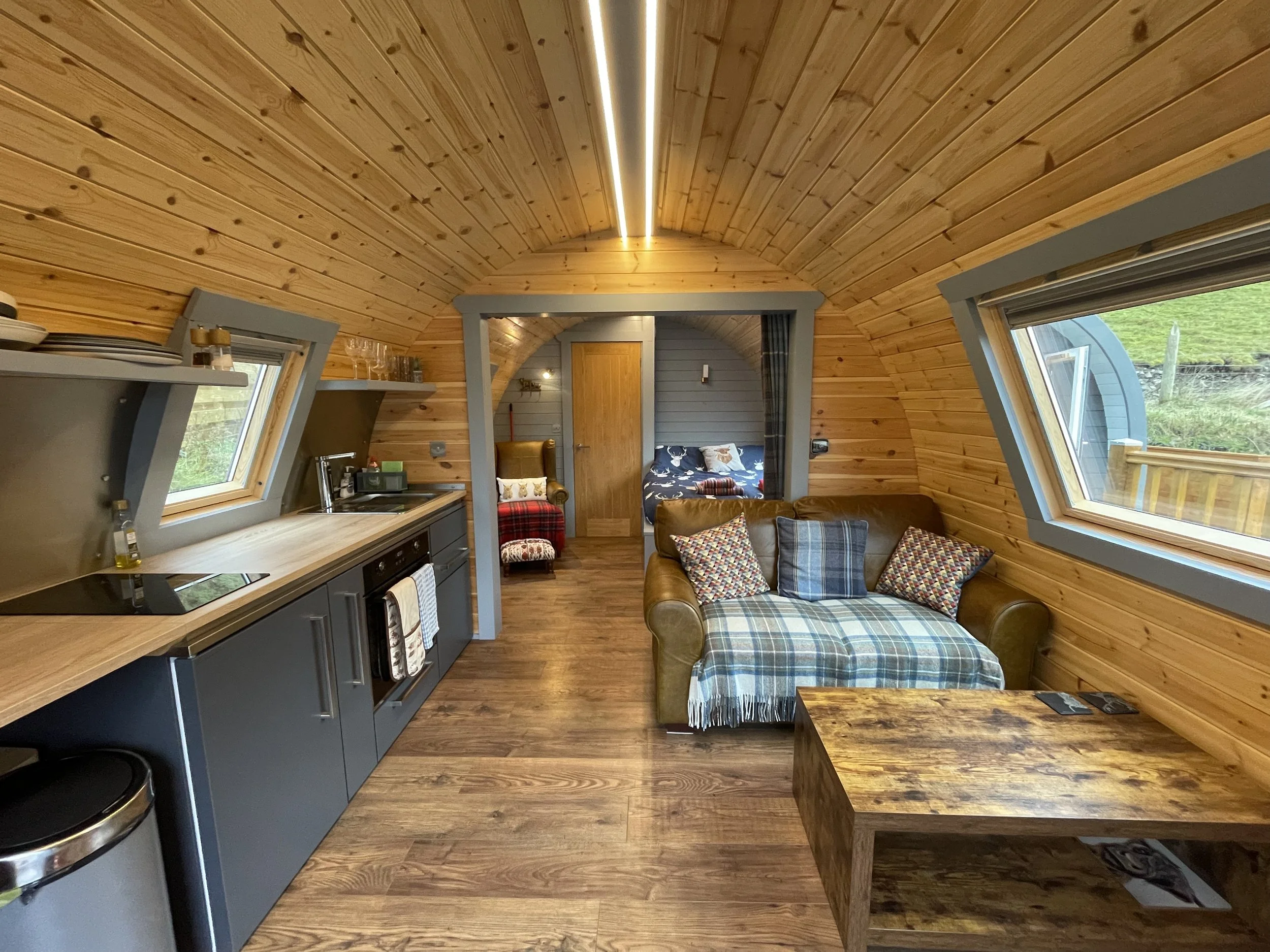 Interior of a cozy wooden cabin with a sloped ceiling, featuring a small kitchen on the left, a plaid sofa with pillows on the right, a wooden coffee table, and a cozy bedroom with a bed and window in the background.