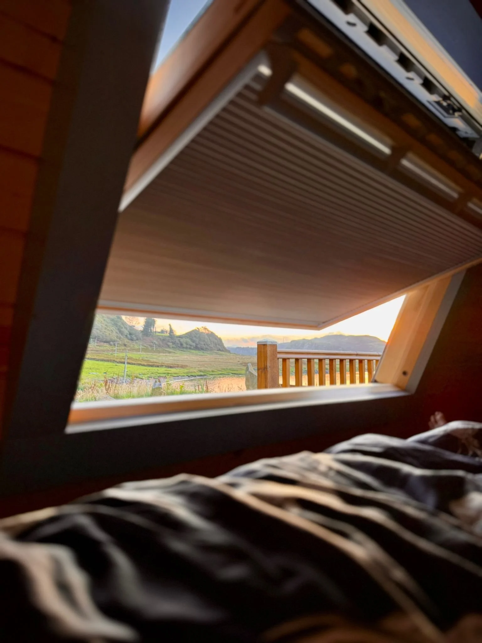View through a tilted window showing a fence, field, trees, and hills at sunset with a blanket in the foreground.