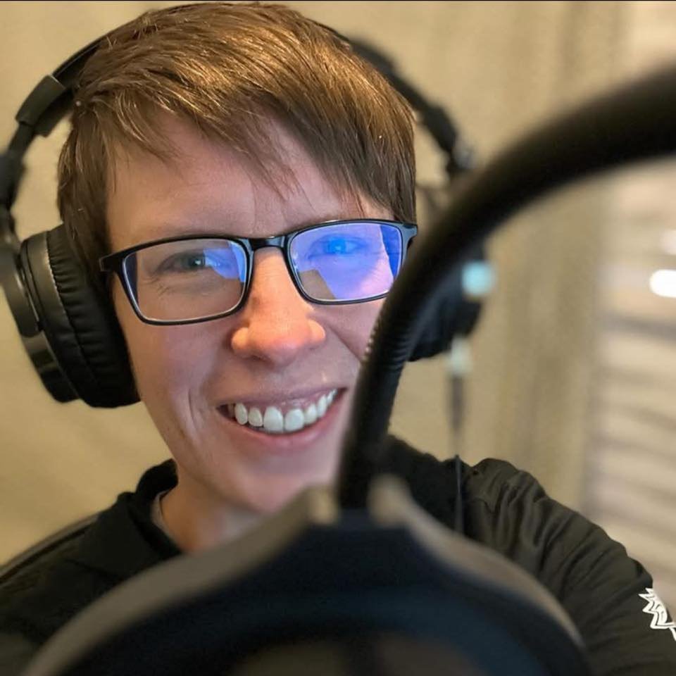 A smiling person with glasses and headphones looking into a camera, with a microphone in front of them.