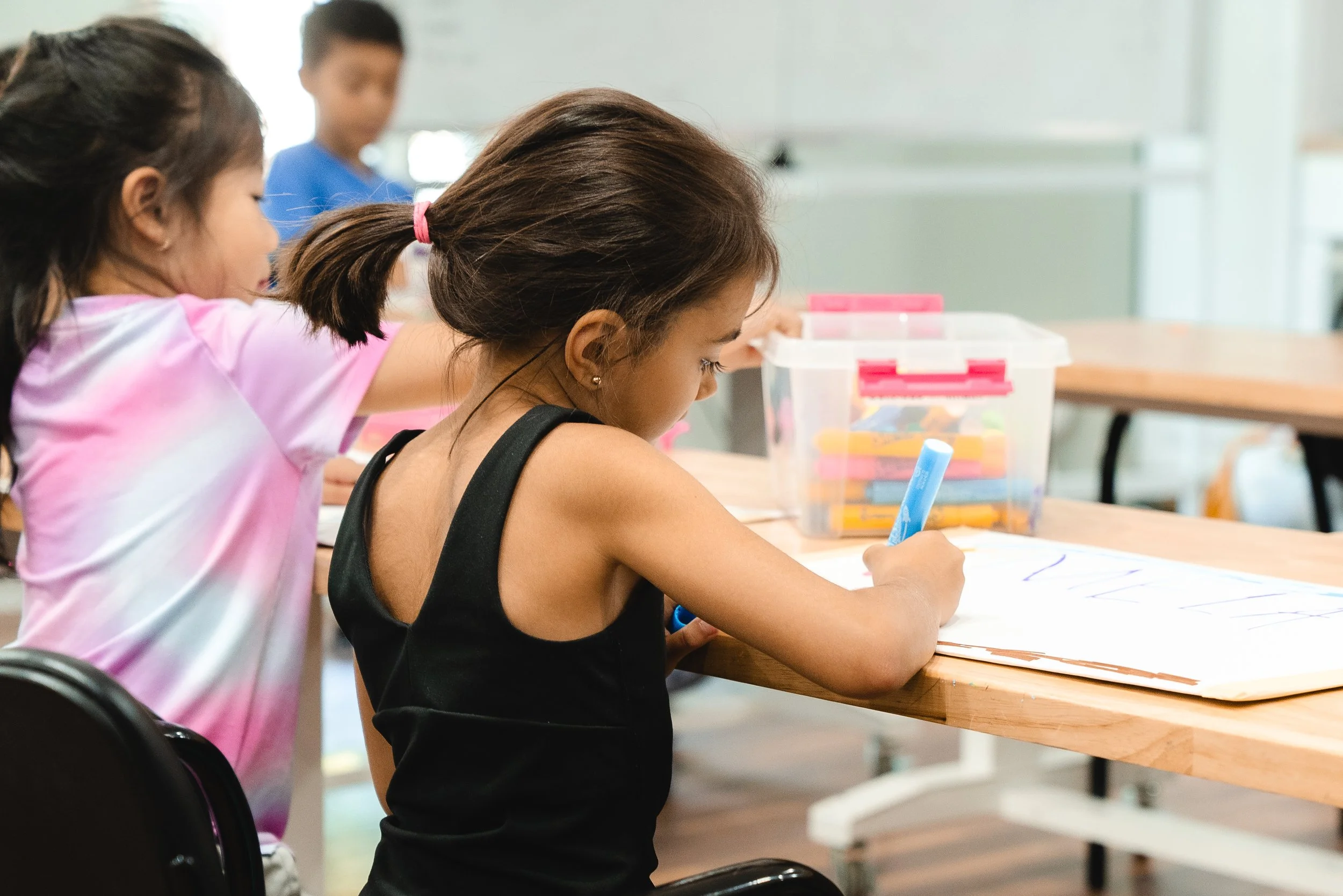 Children sitting at a table, drawing with markers, with a box of markers on the table in a classroom setting.